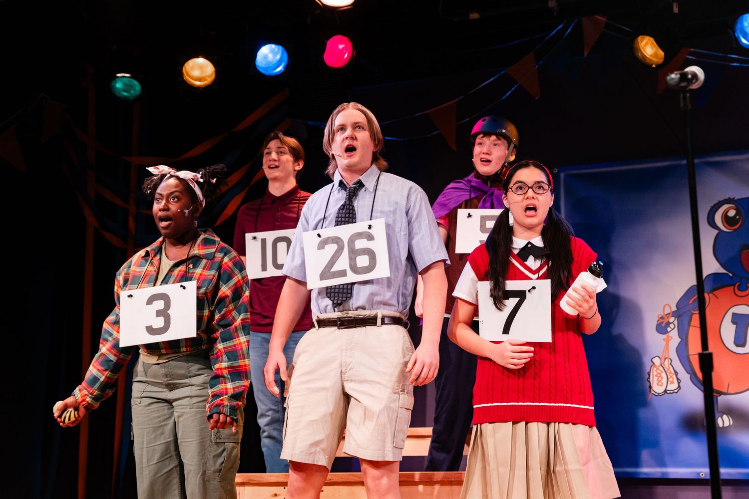 Performers on stage holding numbered signs in a colorful theatrical production under stage lighting.