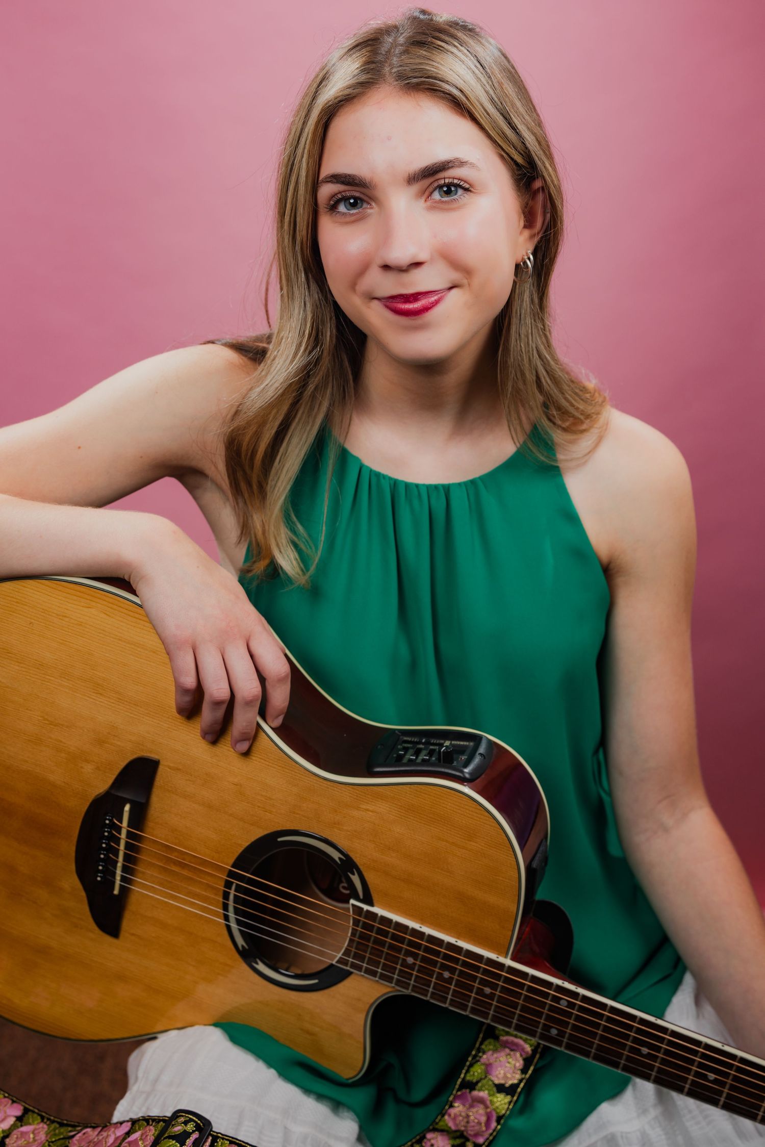 Young blonde woman in green dress holding an acoustic guitar against a pink background.