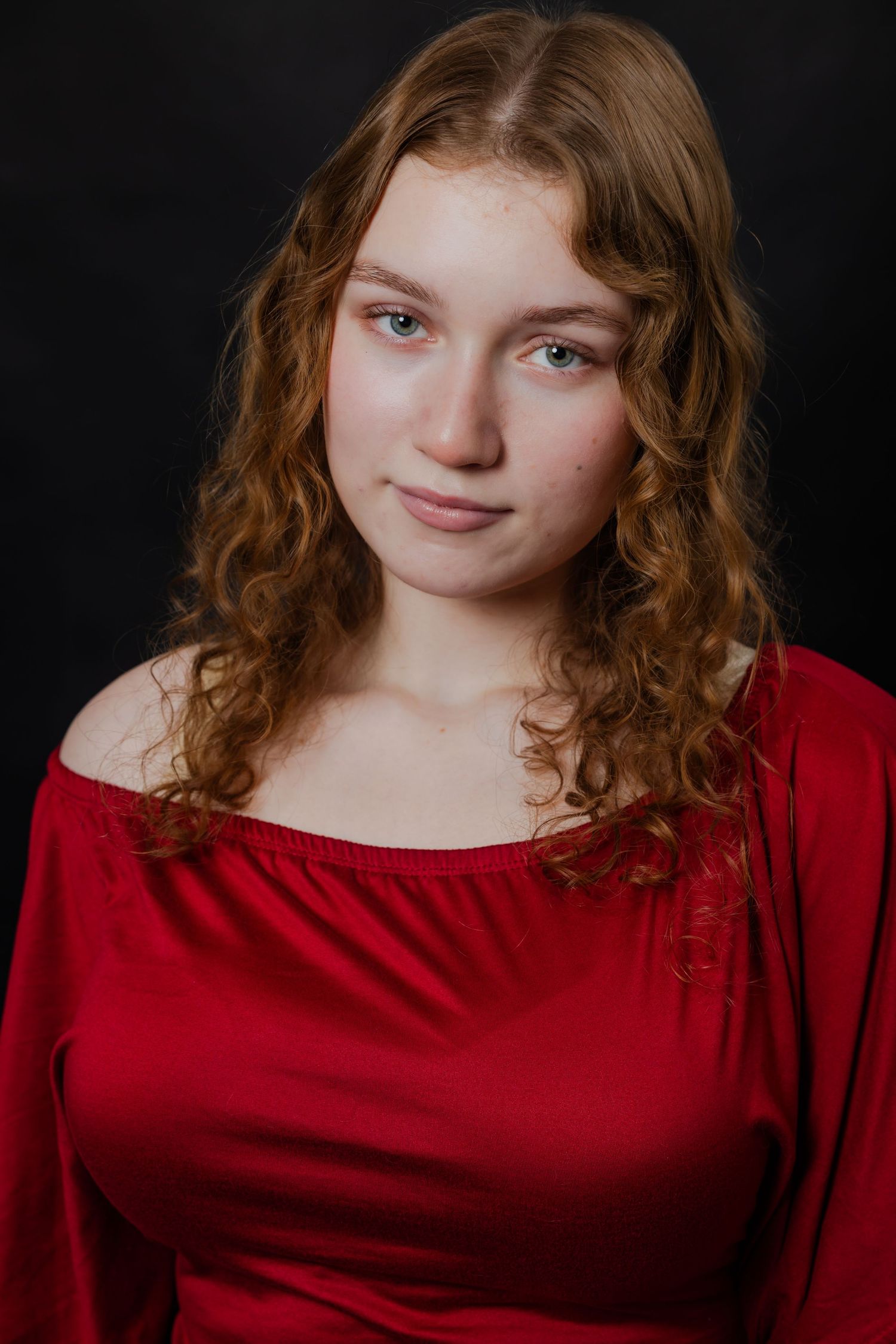 A young woman with curly auburn hair wearing a red off-shoulder top poses against a black background.