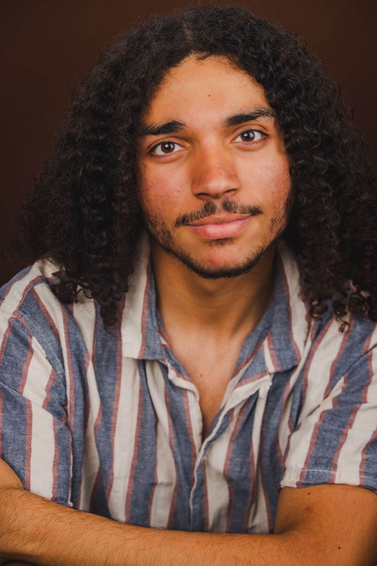 Young man with long curly dark hair and light beard wearing striped shirt, portrait photo.