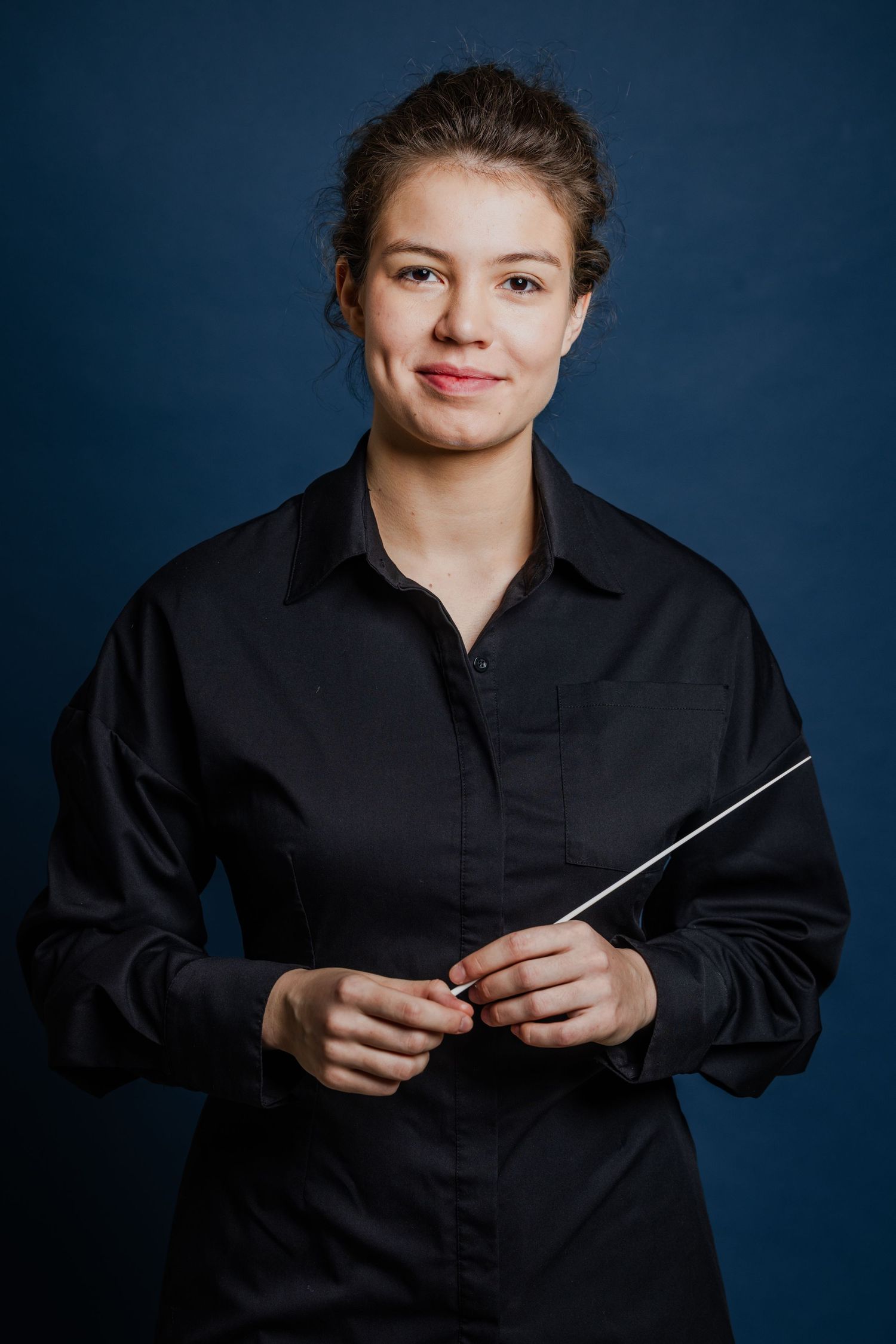 Young female orchestra conductor in black shirt holding a baton, posing against a dark navy blue background.