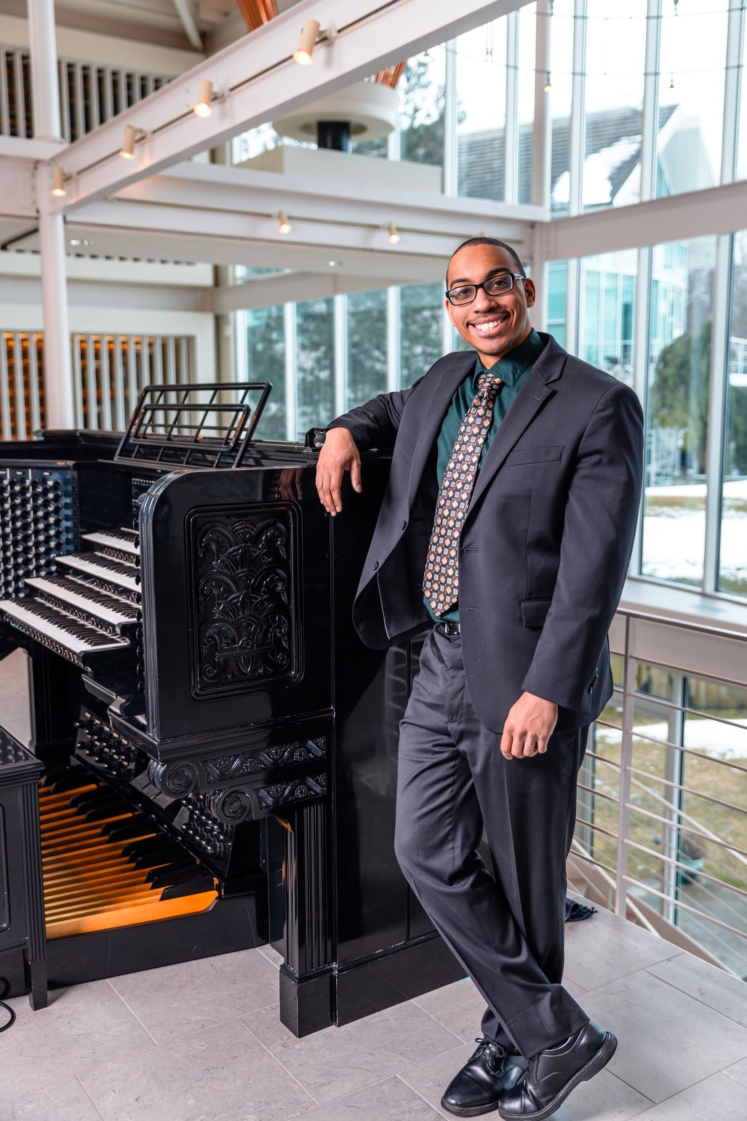 Smiling man in suit leaning against a pipe organ in a bright modern building.