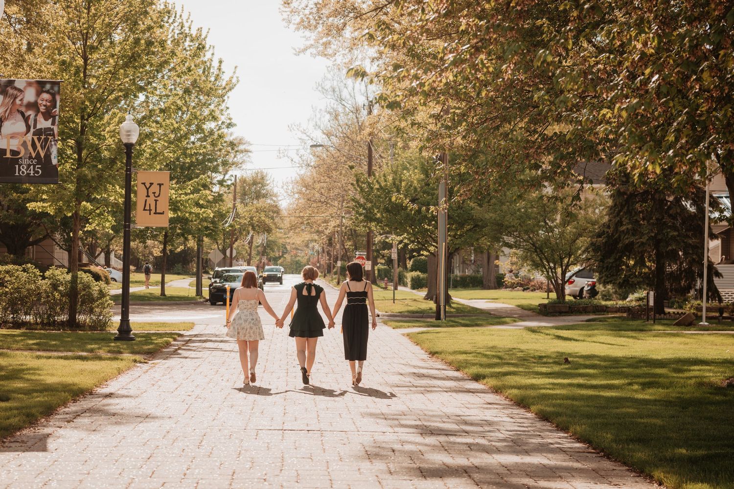 Three women walking hand-in-hand down a tree-lined campus pathway in autumn sunlight.