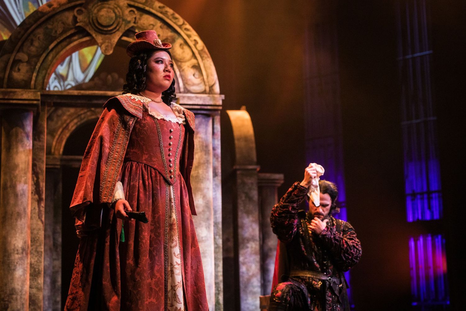 Two performers in Renaissance costumes on a dramatic opera stage with colorful architectural backdrop.