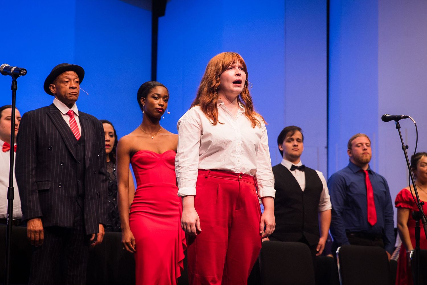 A red-haired soprano soloist performs passionately with a choir on a blue-lit stage.