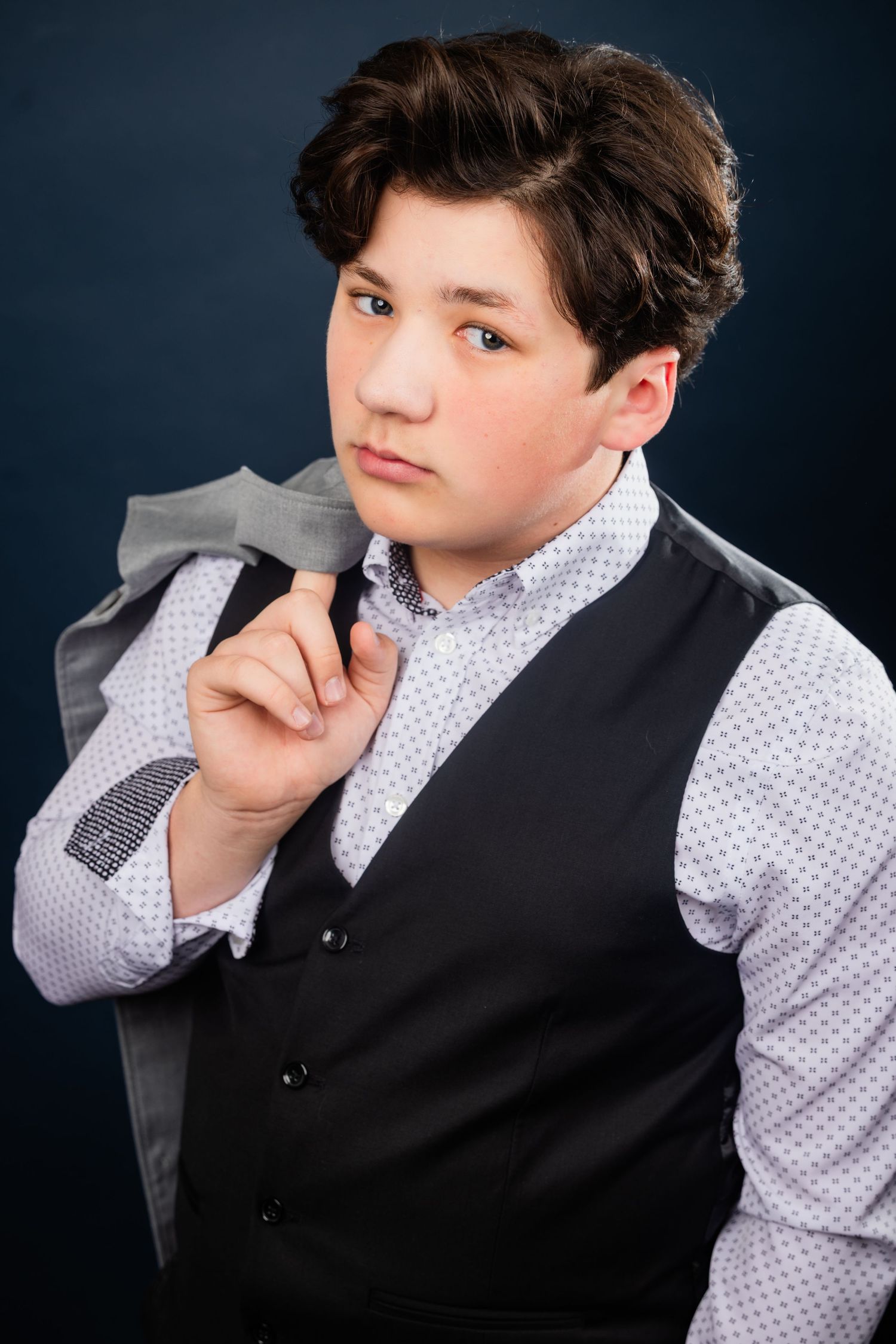 Young boy in a black vest and patterned shirt holding a jacket over his shoulder on dark background.