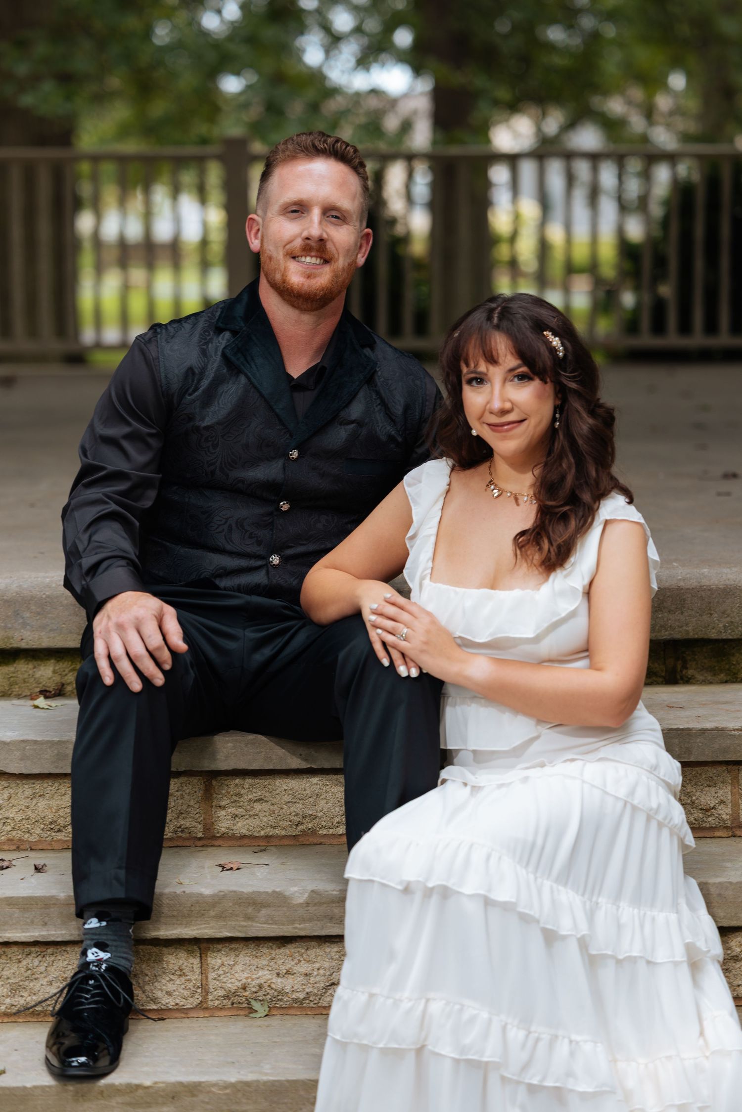 A bride in white ruffled dress and groom in black suit sitting together on stone steps outdoors.