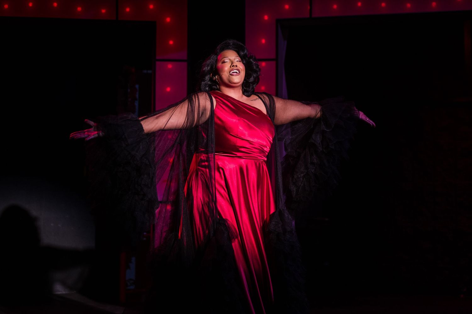 A performer in a red gown with feathered sleeves delivers a powerful solo under dramatic stage lighting.