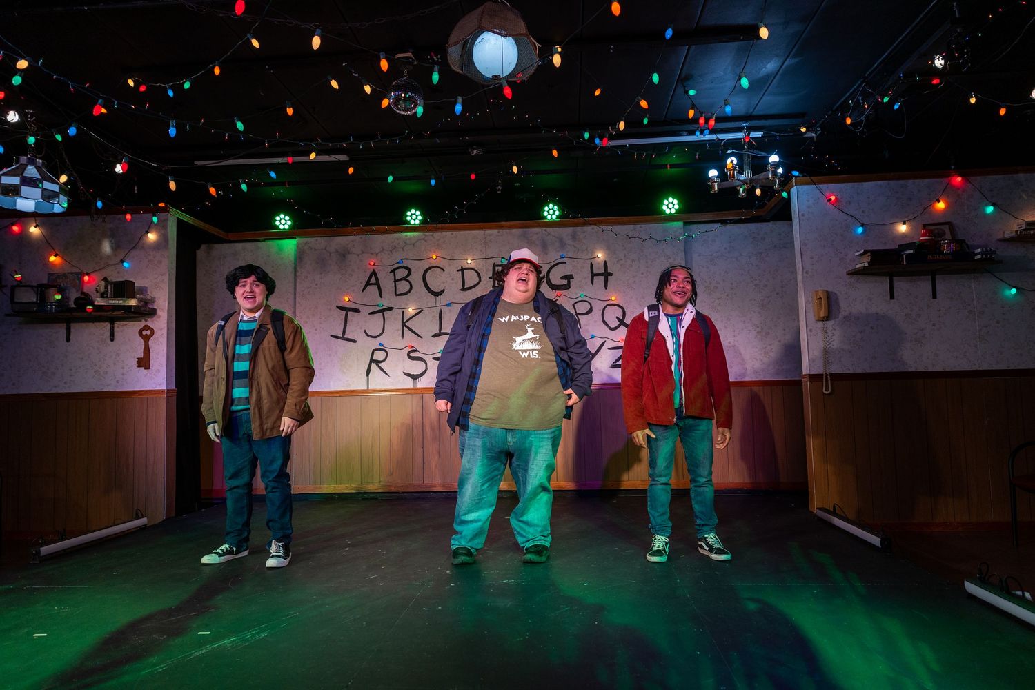 Three young actors performing on stage with a colorful light display and alphabet wall in a Stranger Things parody.