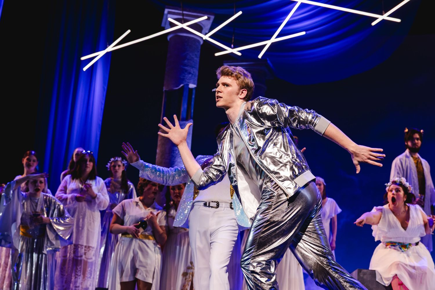 A performer in a silver jacket dances on stage surrounded by ensemble cast members in white costumes.