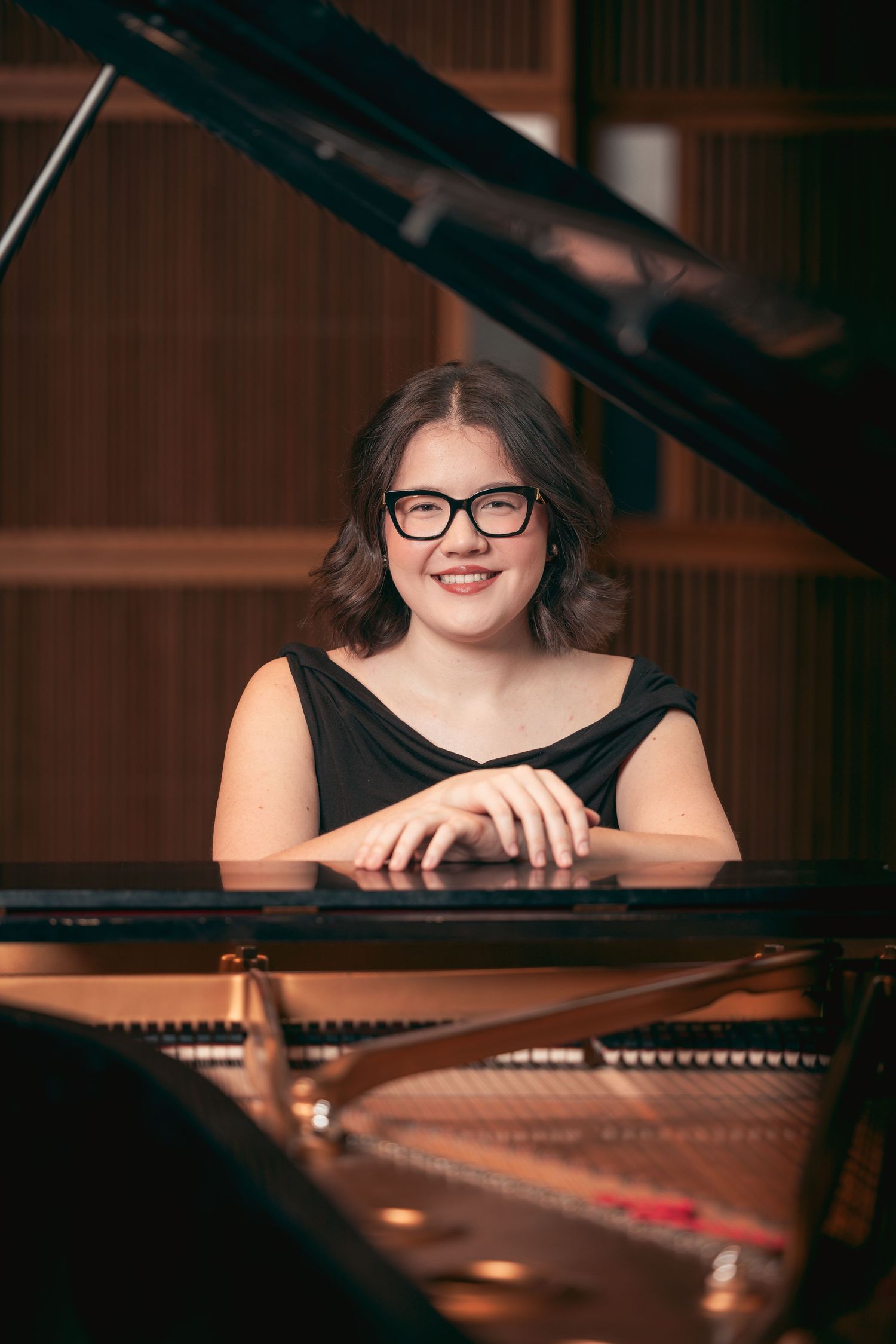 Smiling young woman with glasses seated at a grand piano in a music studio.