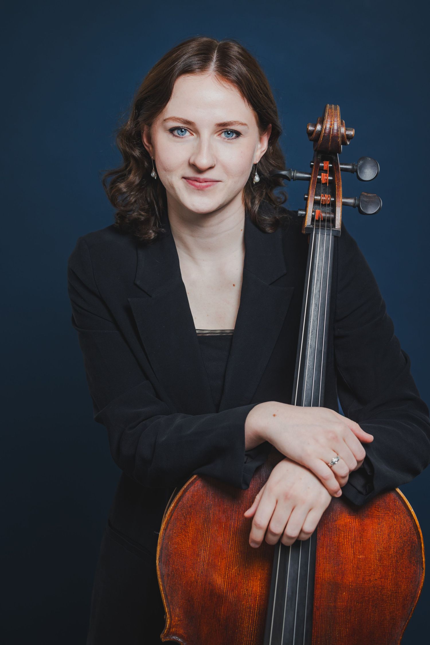 Female cellist in a black blazer leaning on her cello, posing against a dark blue background.