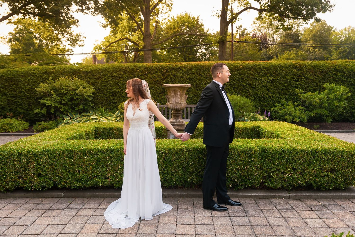 Bride and groom holding hands back-to-back in elegant garden with manicured hedges at sunset.