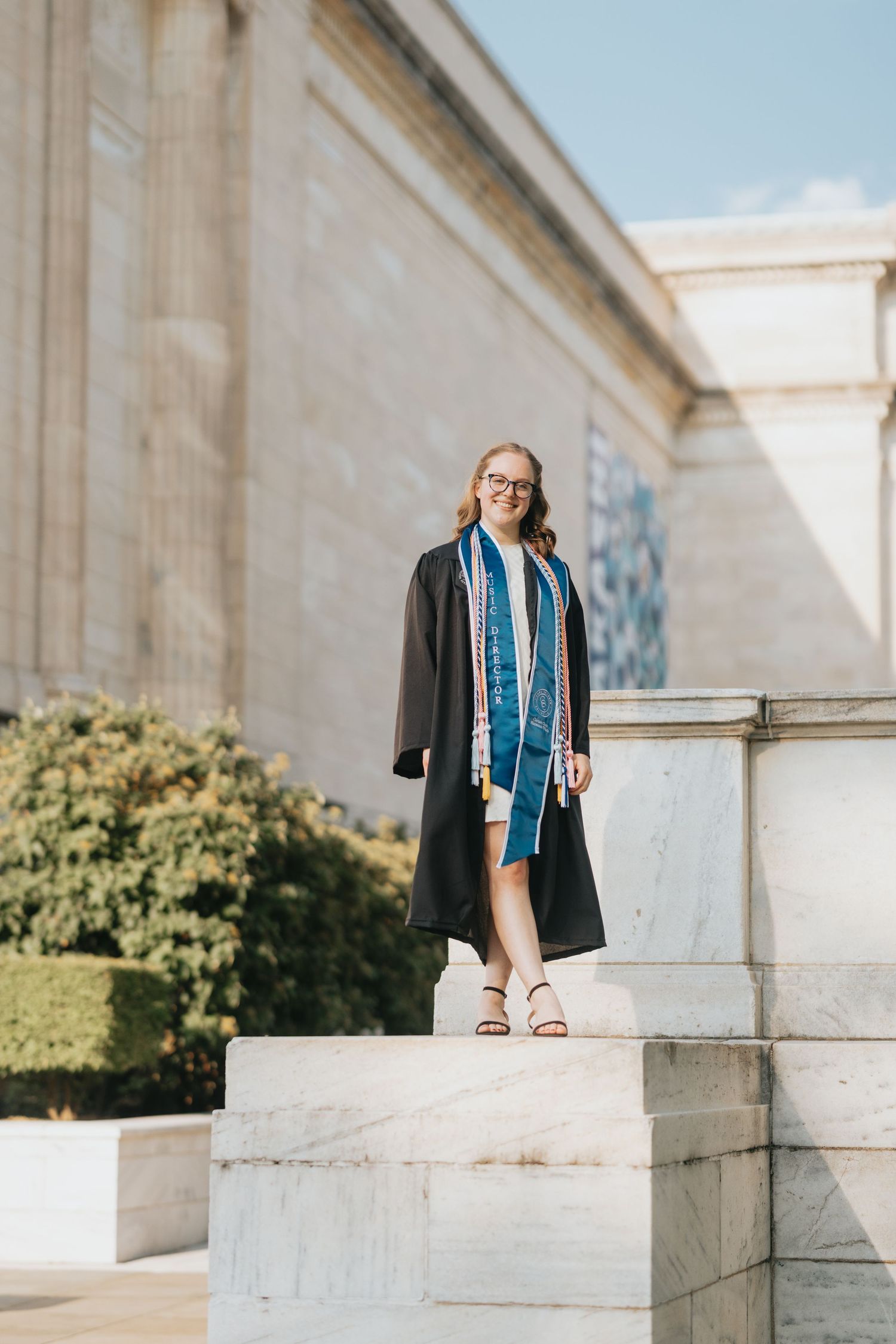 Young woman in graduation gown and stole standing on marble steps of a neoclassical building.