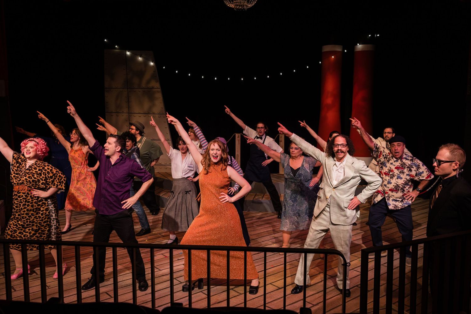 Theater cast performing a choreographed dance number with arms raised on a lit stage.
