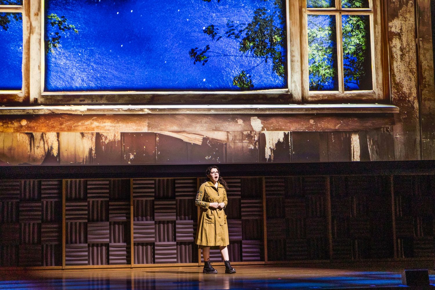 Performer in a tan trench coat stands alone on a theatrical stage with a blue scenic backdrop.