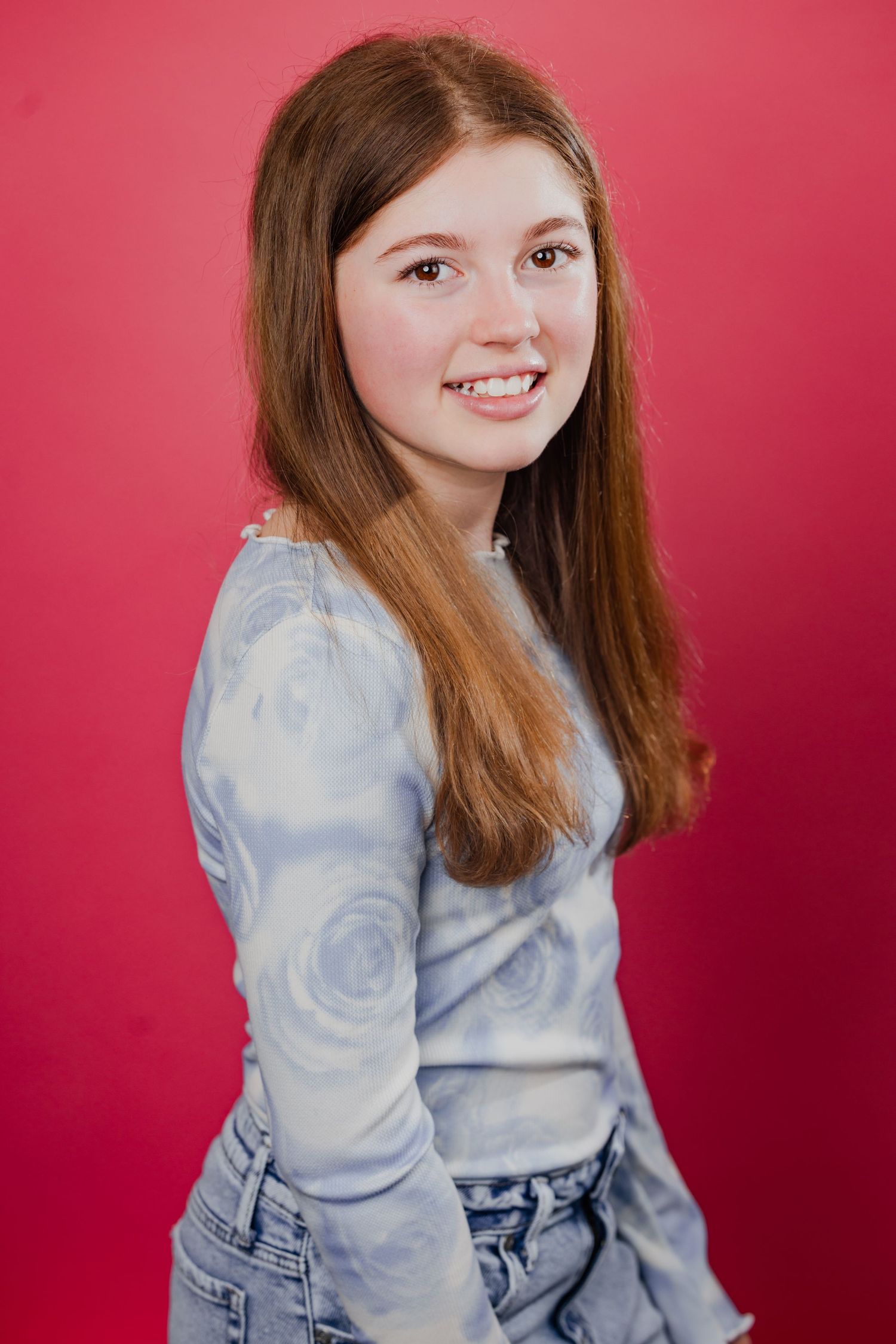 Smiling teenage girl with auburn hair wearing a floral long-sleeve shirt and jeans poses against a red background.