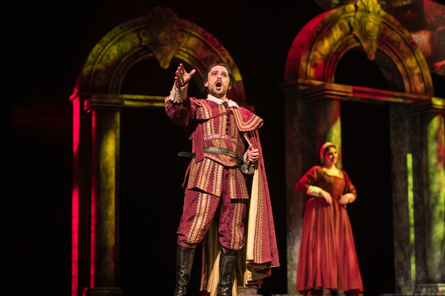 Male opera singer in red Renaissance costume performing on stage with a woman in red dress behind him.