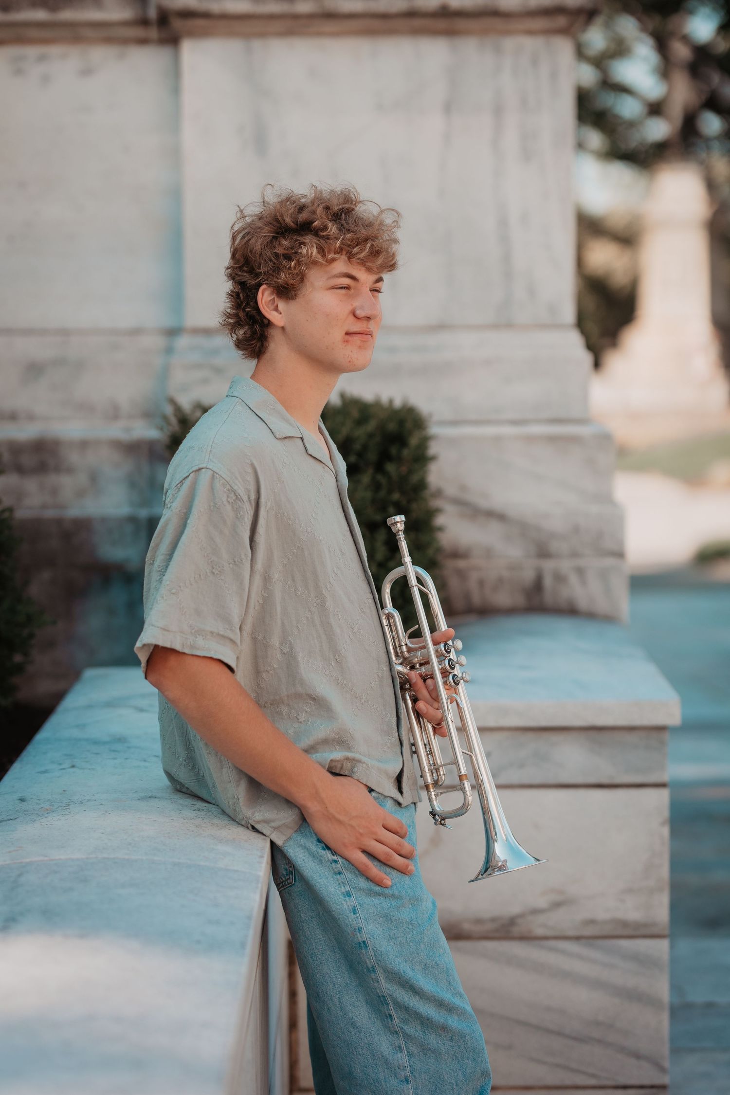 Young man with curly hair holding a silver trumpet while leaning against a marble wall outdoors.