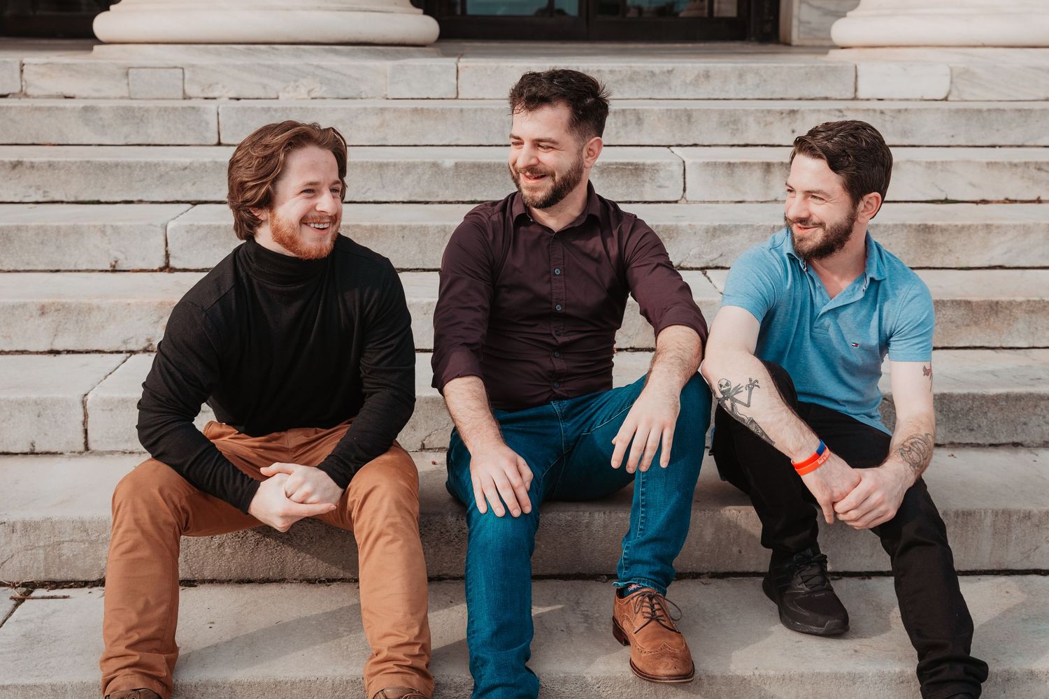 Three young men sitting on stone steps laughing and chatting in casual outfits.