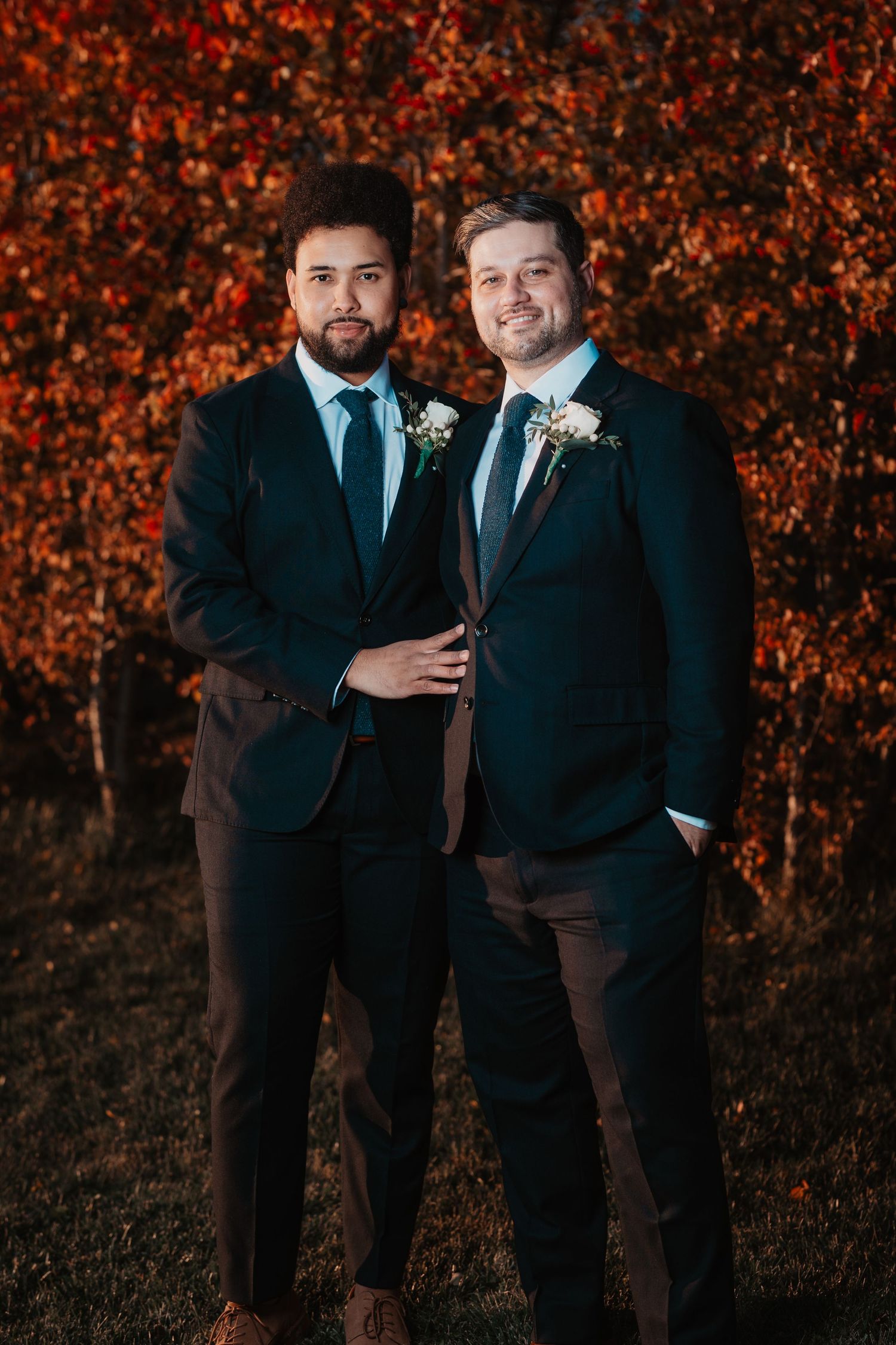 Two men in dark suits with boutonnieres pose before vibrant red autumn foliage.