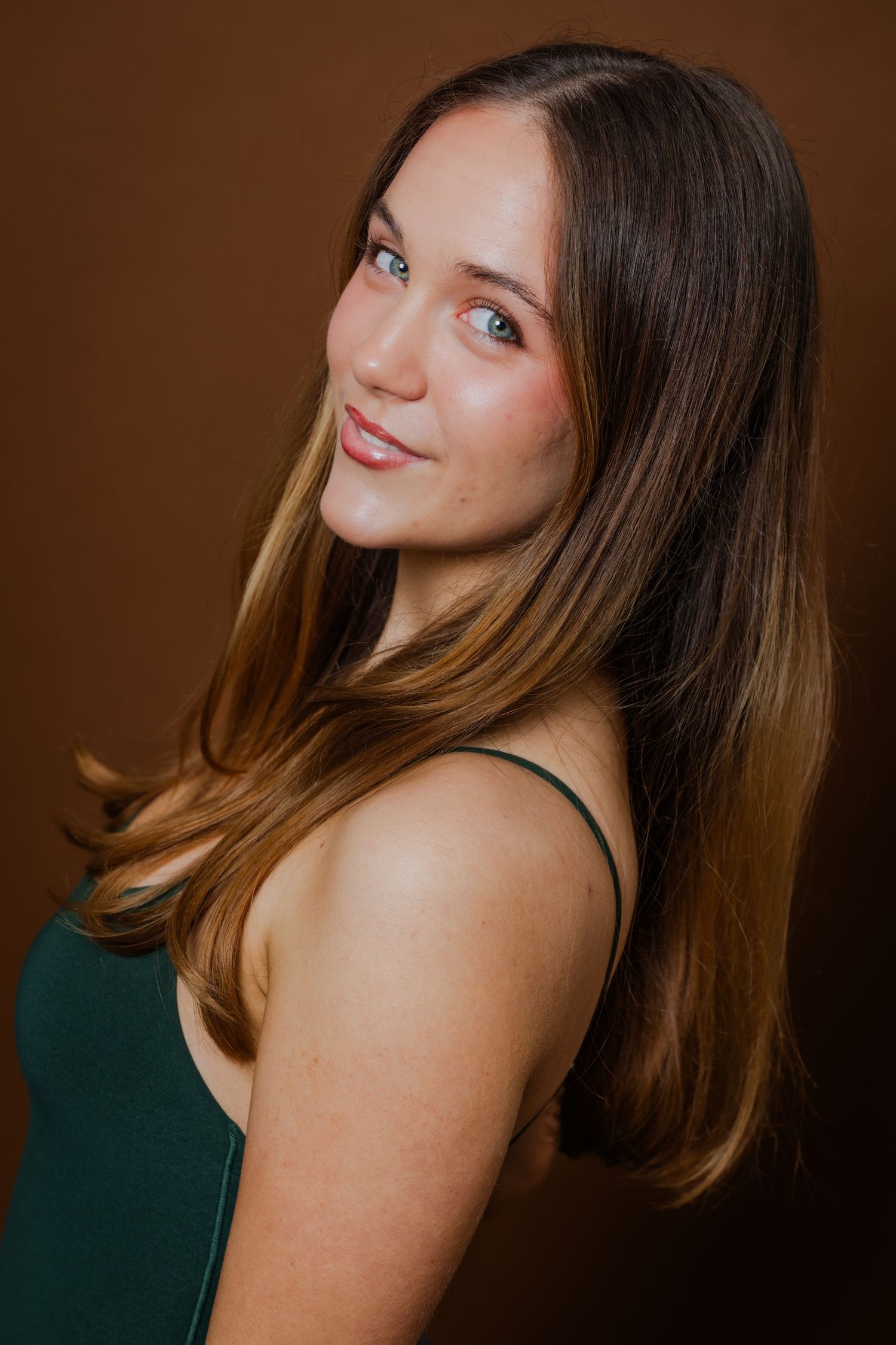 Smiling young woman with long brown hair and blue eyes wearing a green top against a brown background.