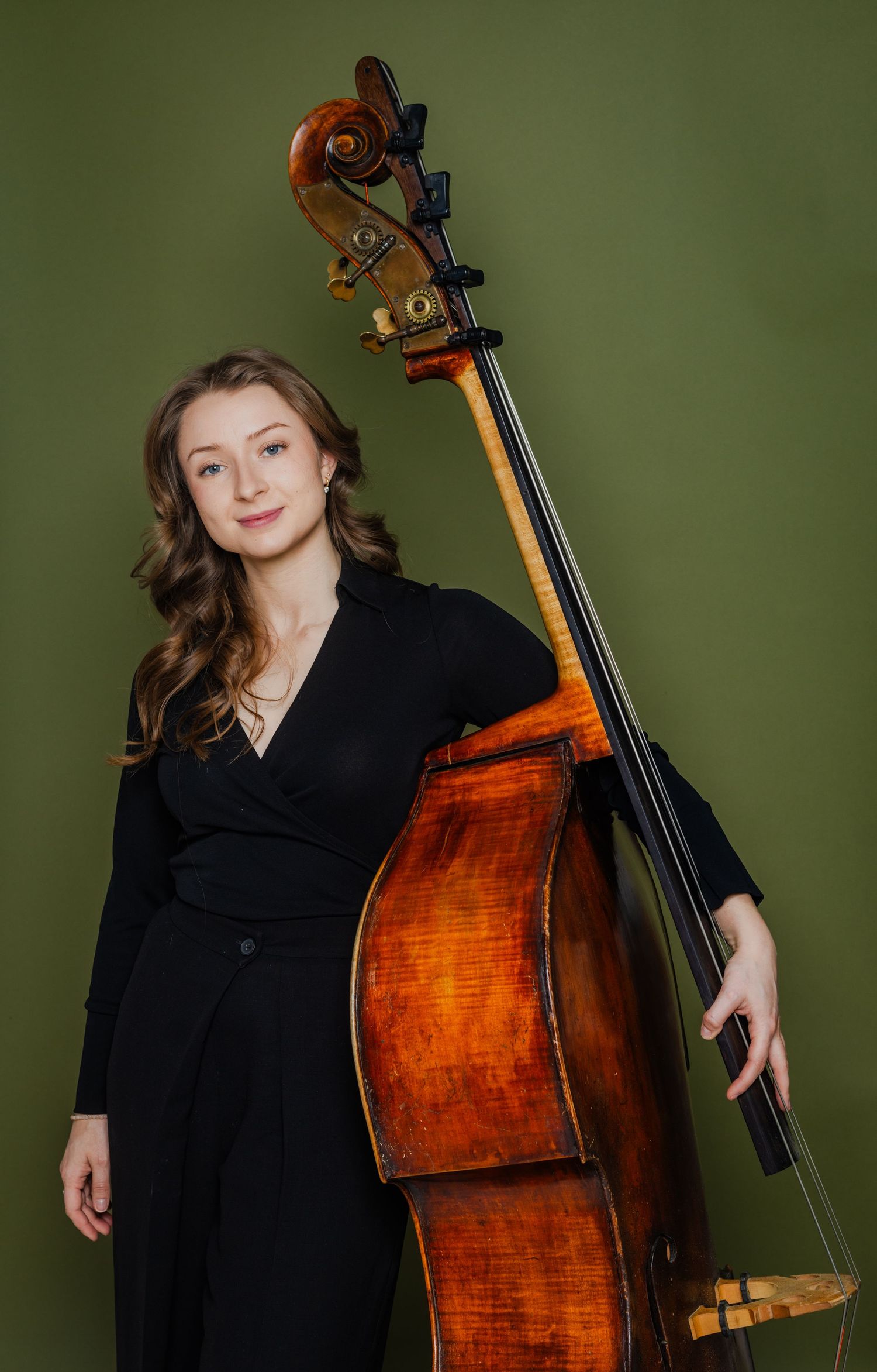 Young woman in black holding a double bass against an olive green background.