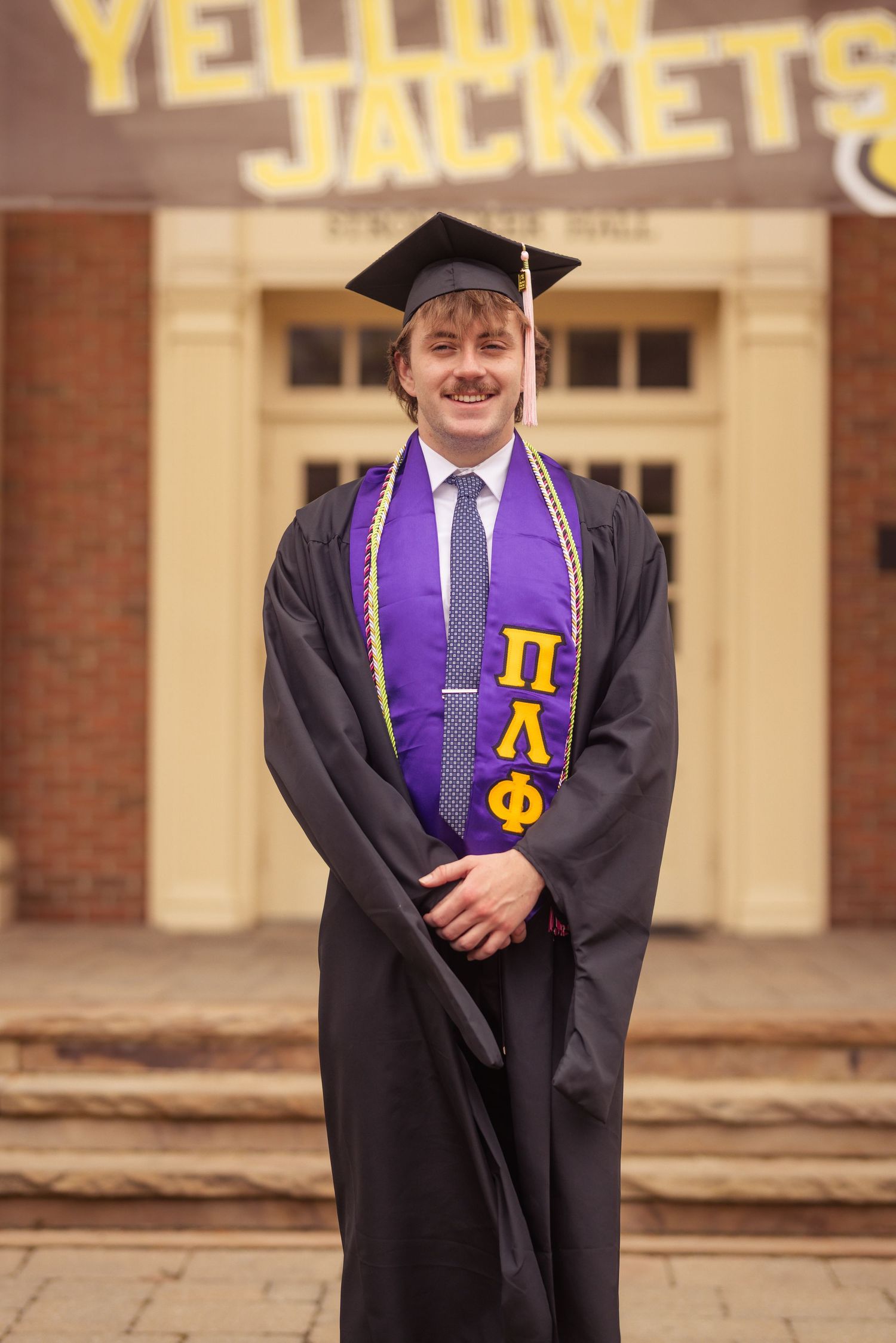 Young man in graduation gown and cap wearing Pi Lambda Phi fraternity stole in front of Yellow Jackets building.