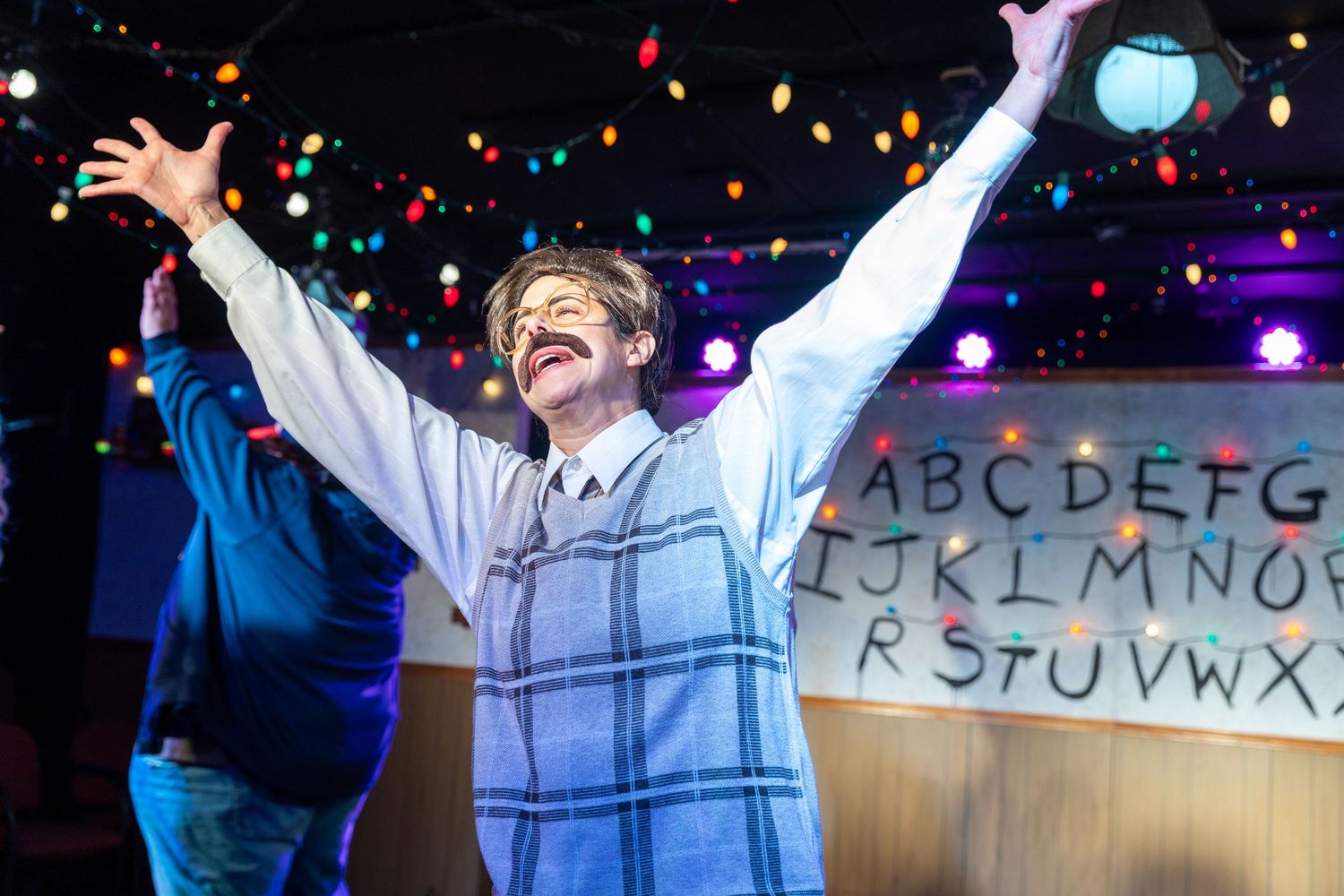 Actor with mustache raises arms triumphantly on stage during a Stranger Things-themed theatrical performance.