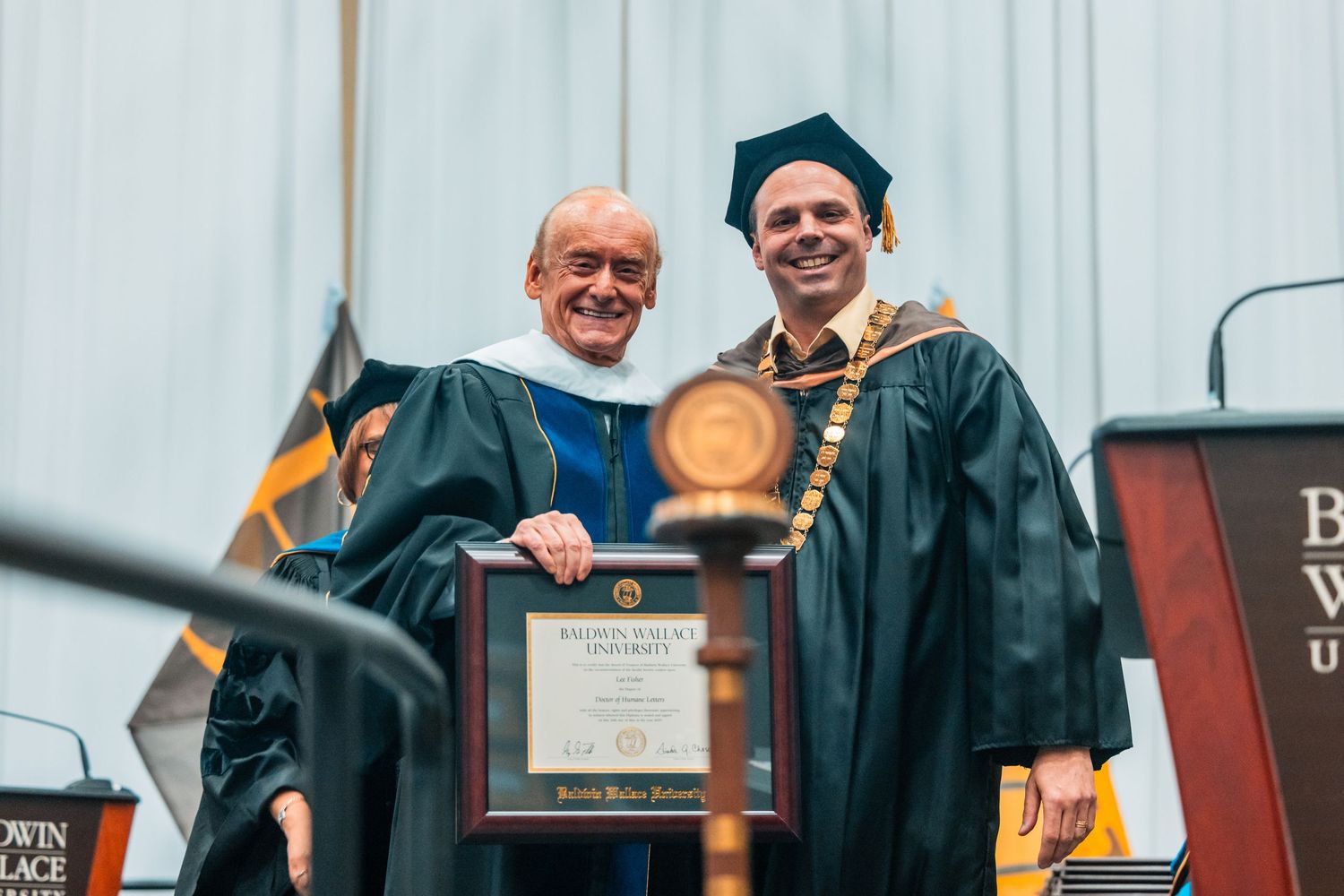 Two men in academic regalia smiling while holding a framed award at a university graduation ceremony.