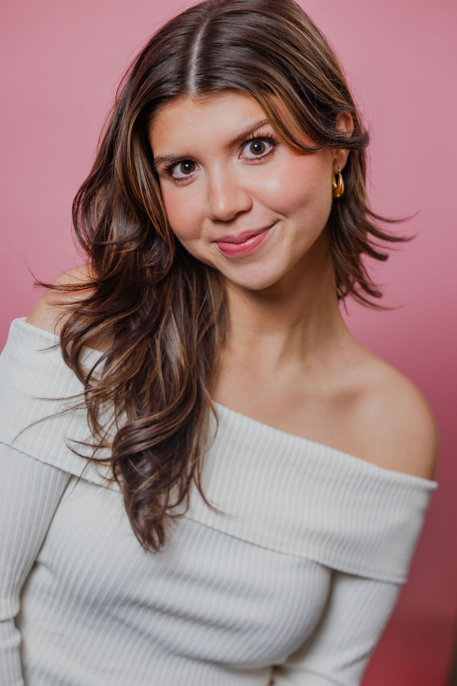 A young woman with highlighted brown hair wearing an off-shoulder white top poses against a pink background.