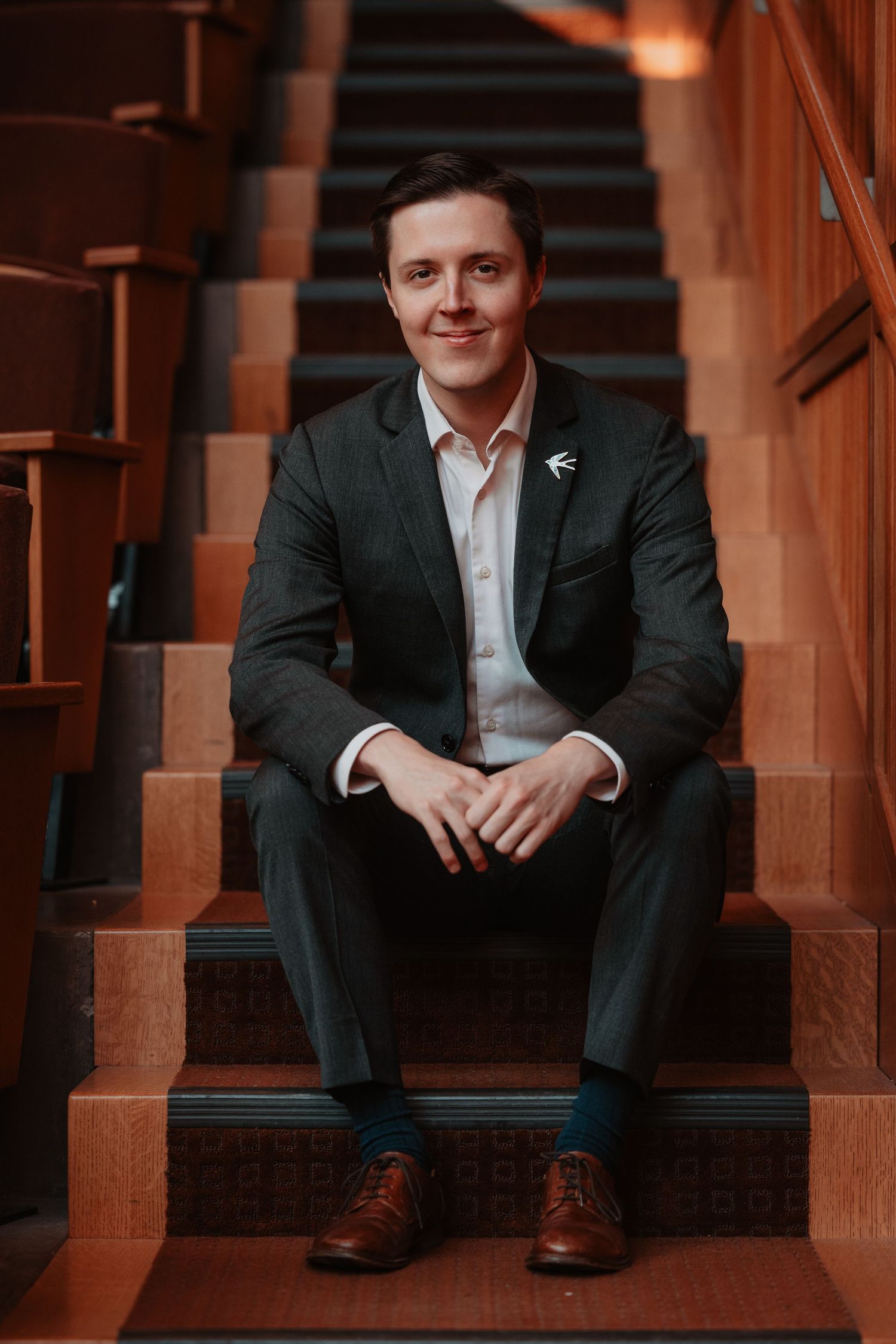 Smiling man in dark suit sitting on wooden staircase indoors.