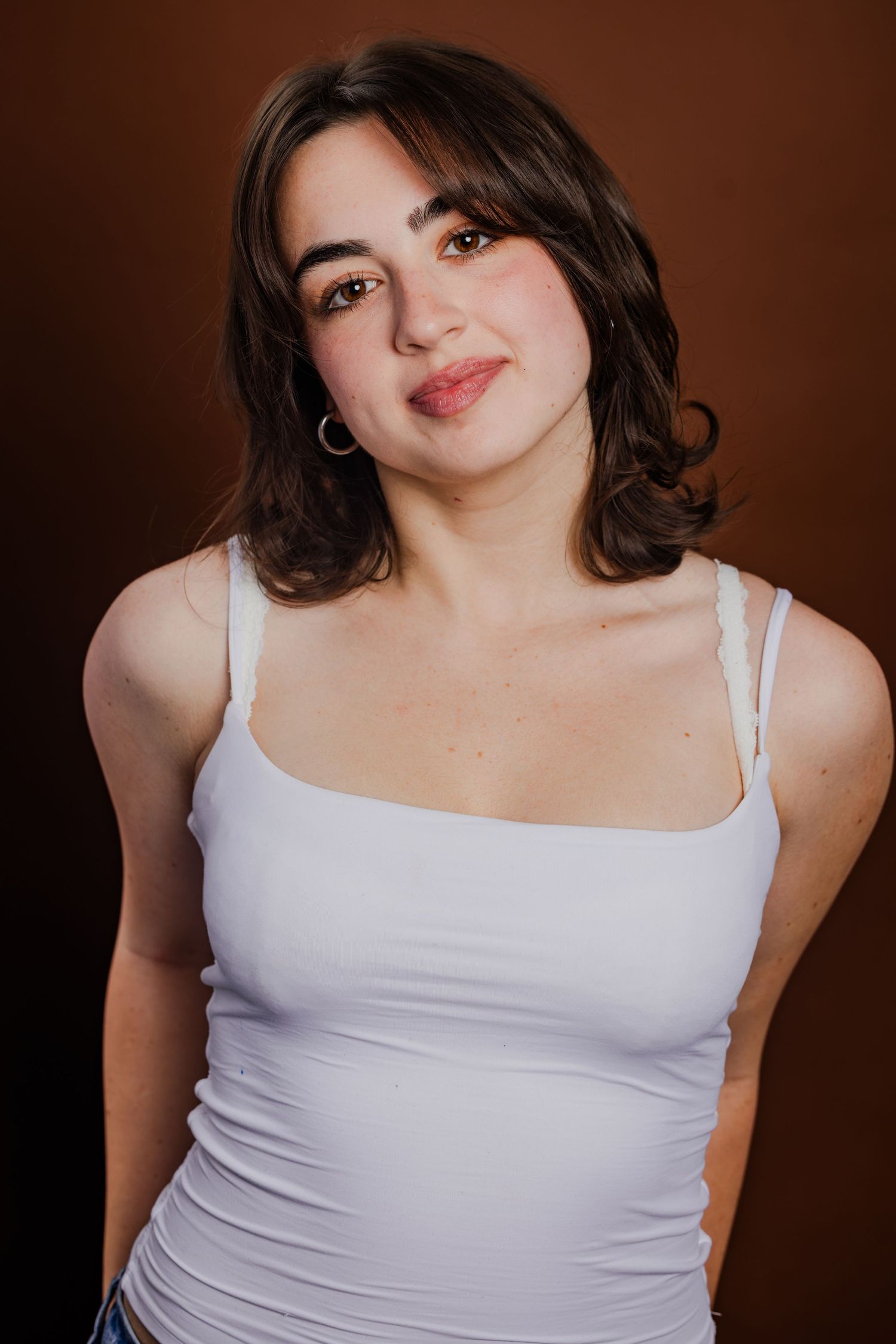 Young woman in white tank top with hoop earrings posing against a warm brown studio background.