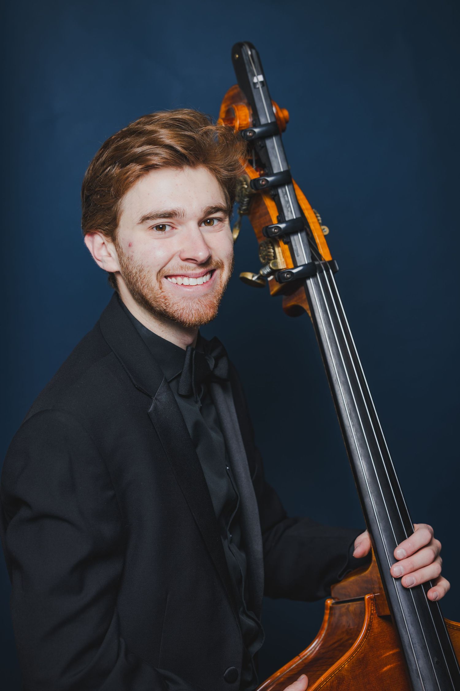 Smiling young male musician with a red beard holding a double bass in a dark blue studio portrait.