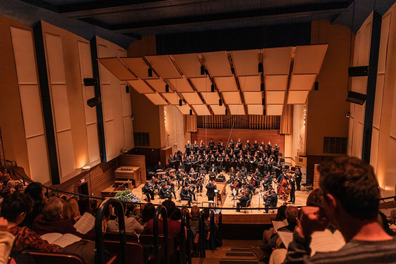 An aerial view of a full orchestra and choir performing in a modern concert hall with wood paneling.
