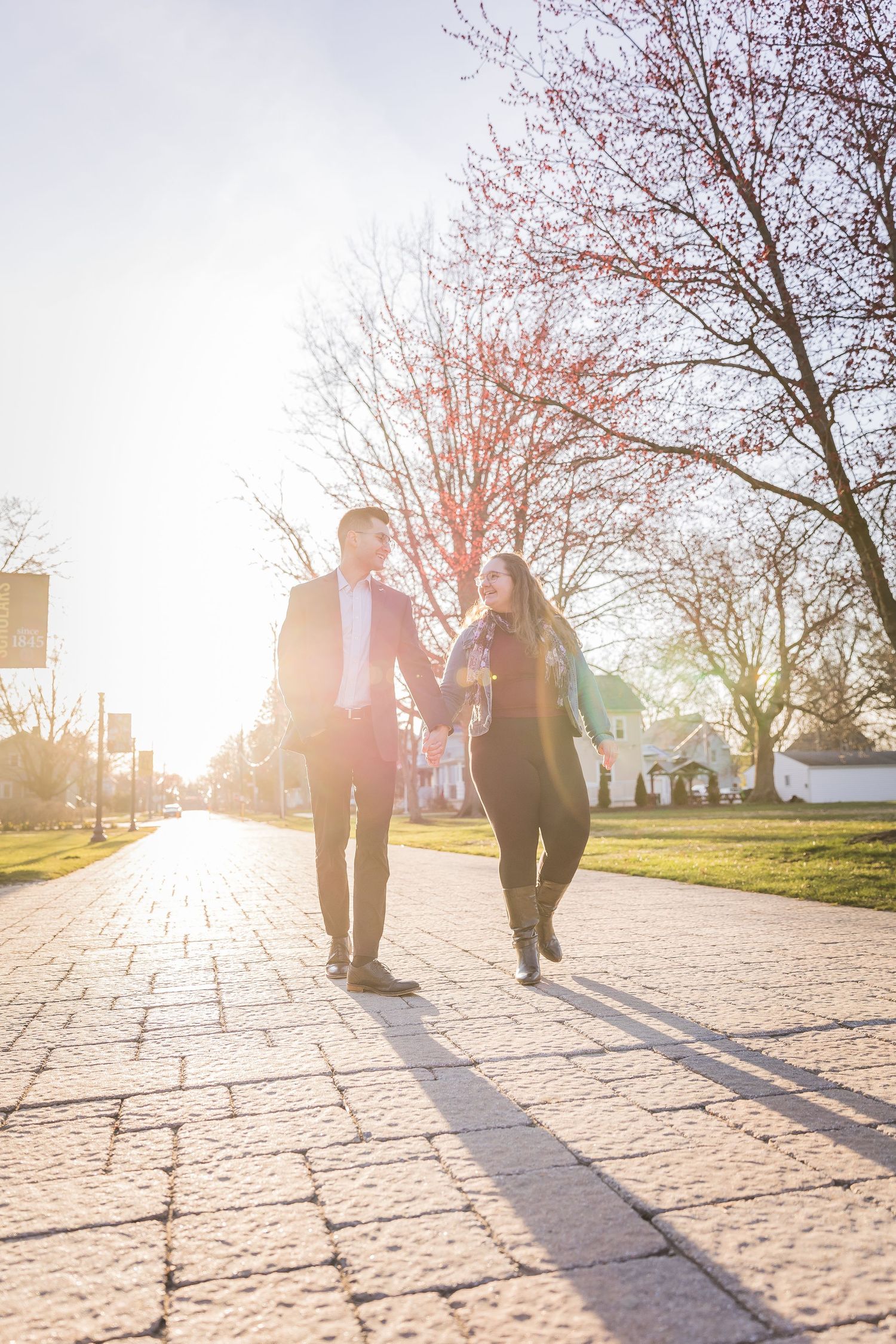 Happy couple holding hands walking on brick path at golden hour with blooming spring trees.