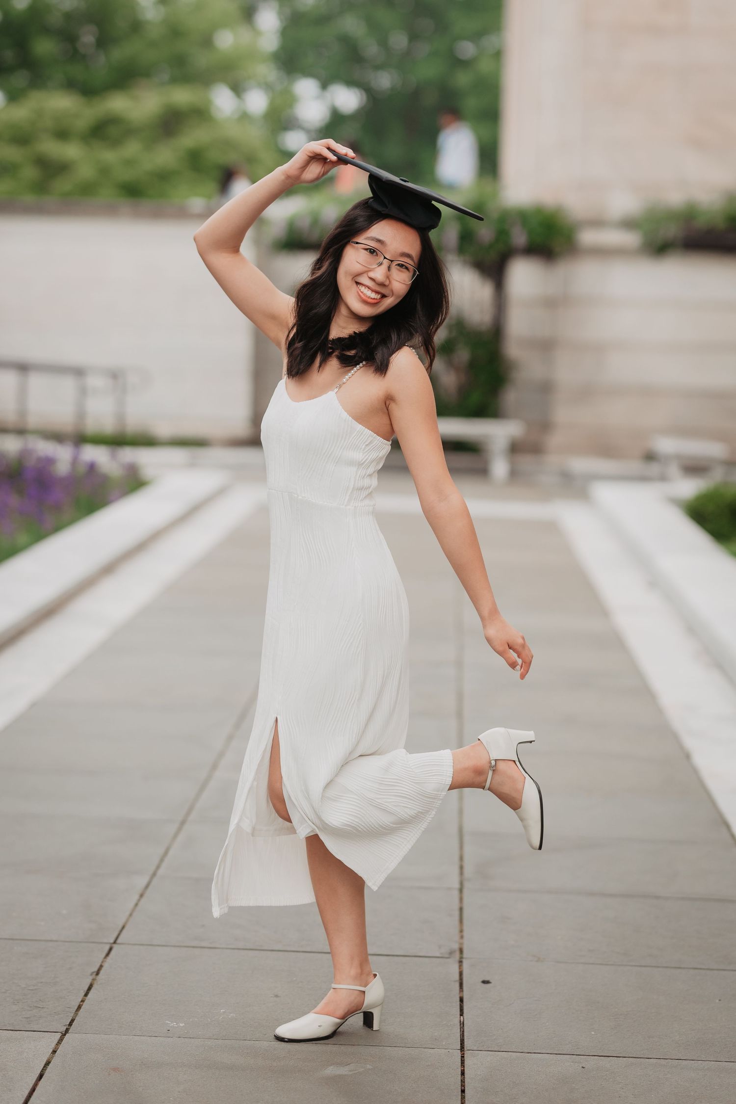 Smiling Asian woman in white dress twirling while holding graduation cap outdoors.