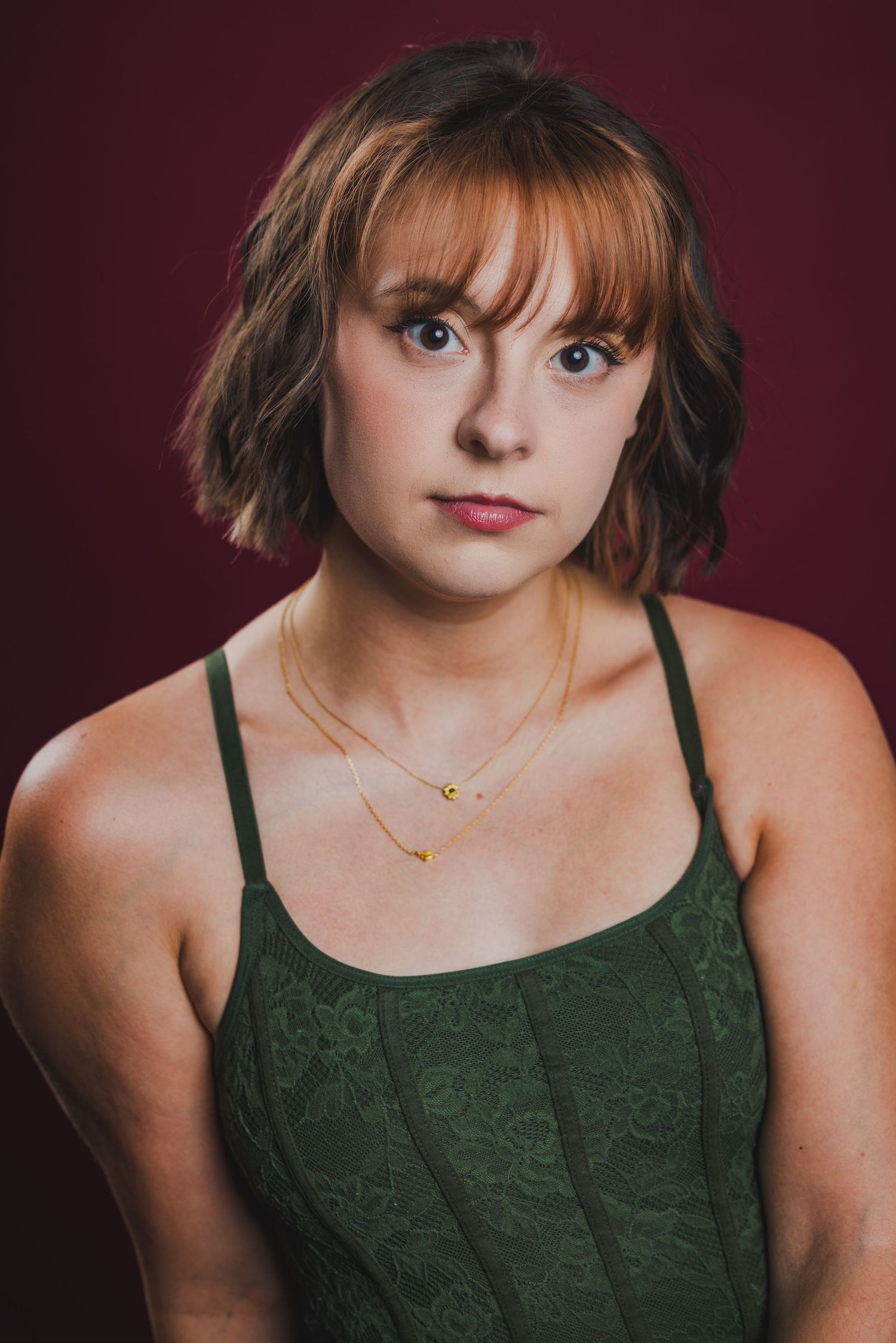Young woman with short auburn hair and bangs wearing green lace top and gold necklaces on burgundy background.
