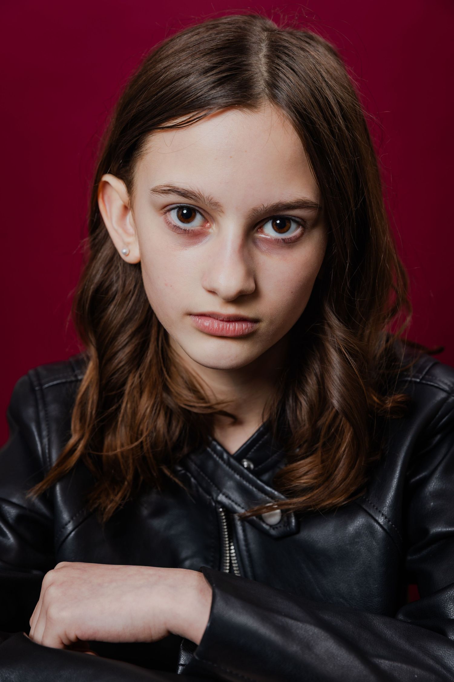 Young girl with wavy brown hair wearing a black leather jacket, posing against a deep red background.