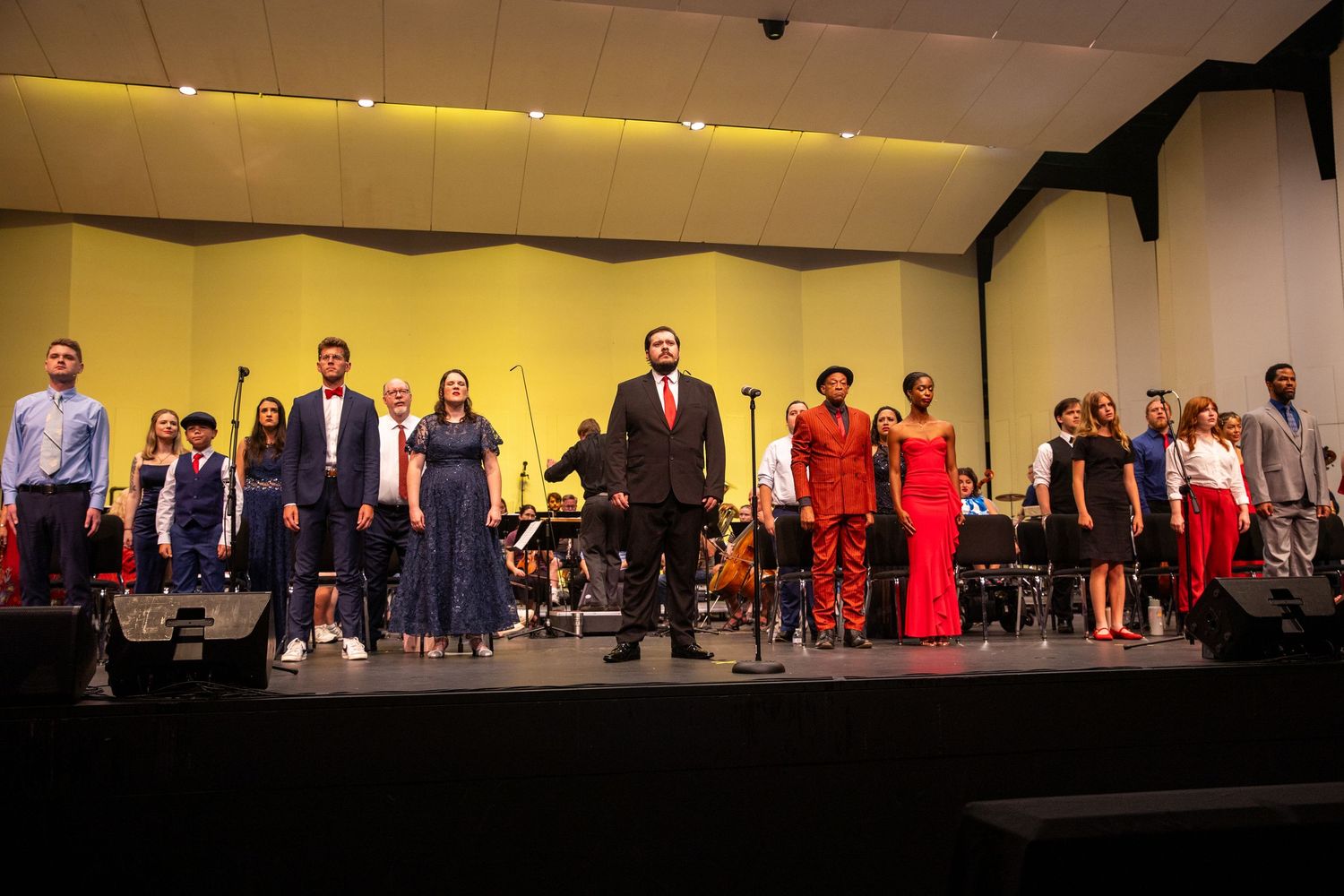 A vocal ensemble and orchestra take a bow on a yellow-lit auditorium stage.