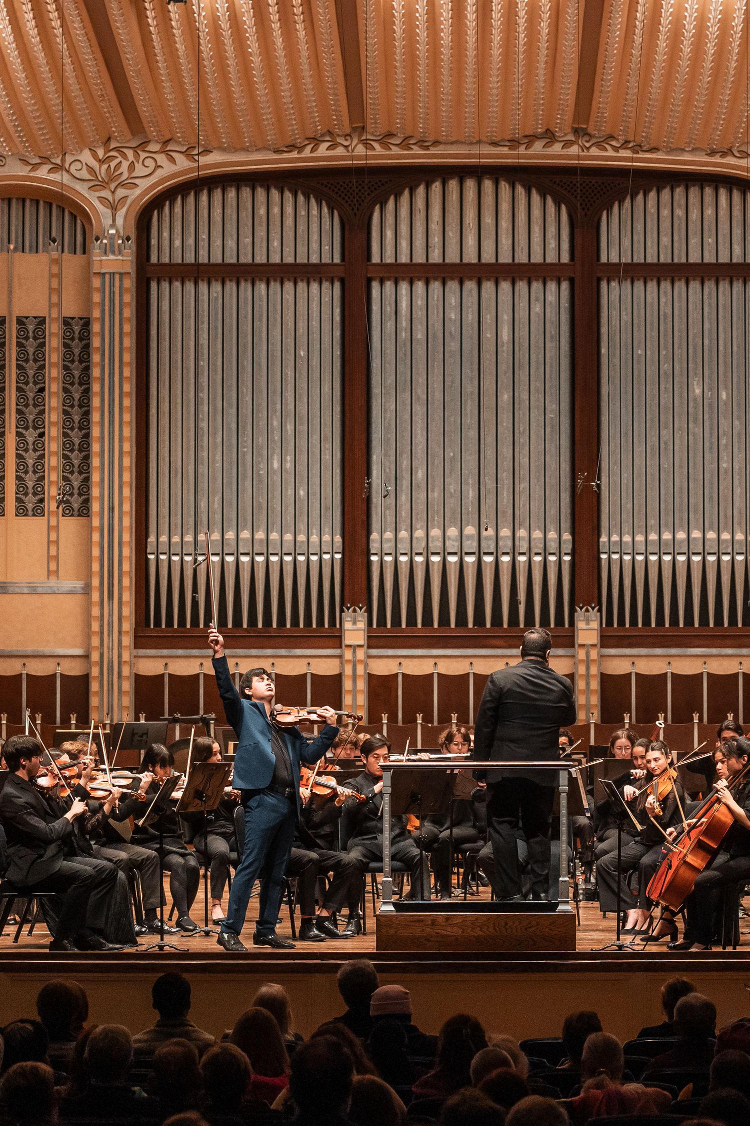 Orchestra conductor and soloist performing on stage in an ornate concert hall with pipe organ backdrop.