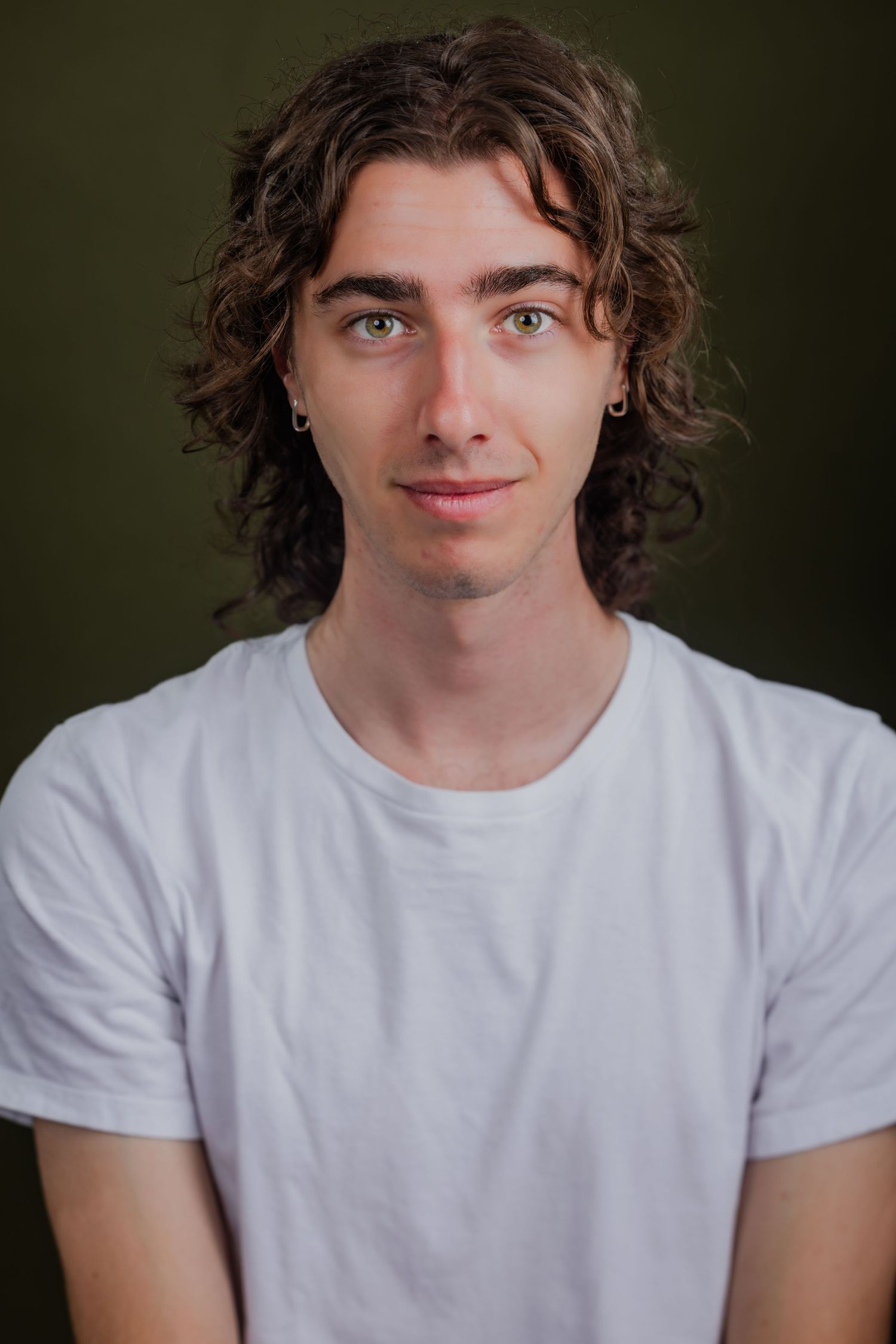Young man with long curly brown hair in a white t-shirt against an olive green background.