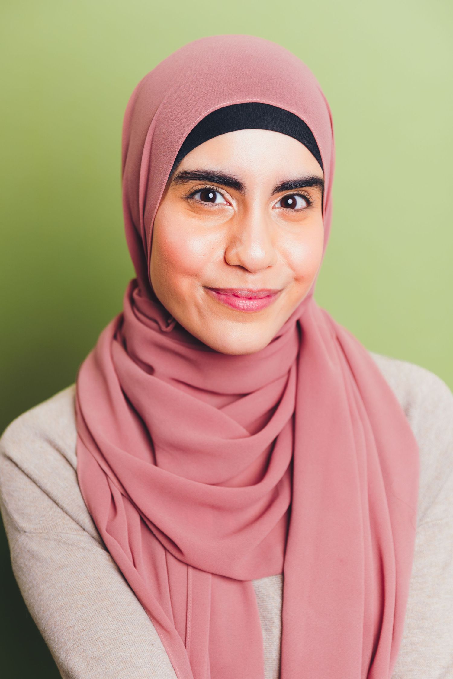 Young woman wearing pink hijab smiling against olive green background in casual portrait.