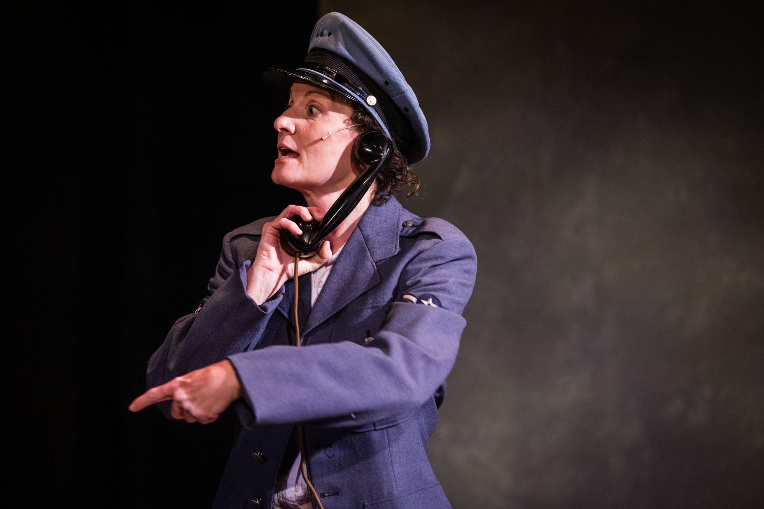Performer in a navy blue uniform and captain's hat gestures dramatically on a theatrical stage.