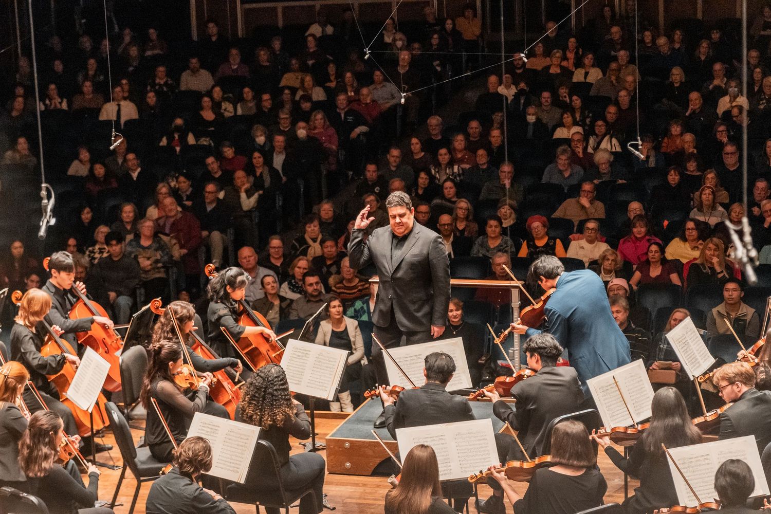 An orchestra conductor leads a full ensemble performance in a packed concert hall with an audience.