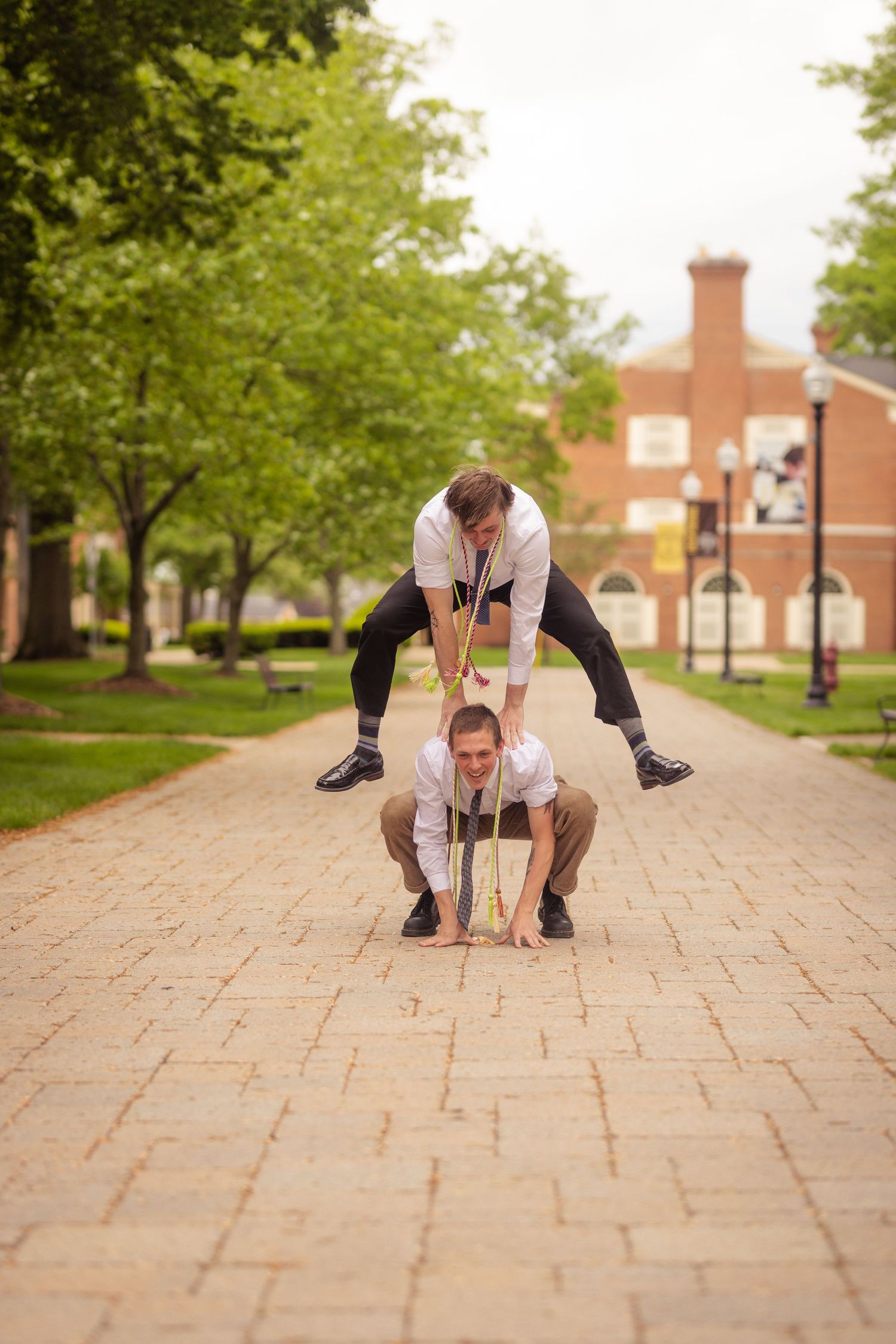 Two young men in graduation attire doing leapfrog on a brick campus pathway.