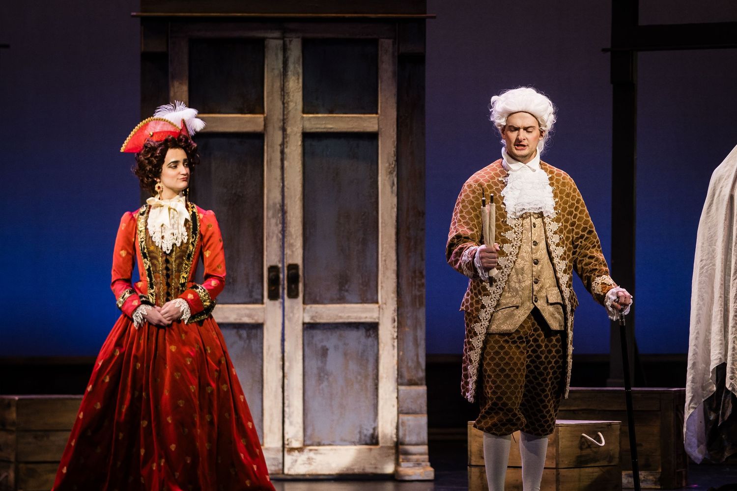 Two performers in period costumes, a woman in red and a man in a powdered wig, perform on a theatrical stage.