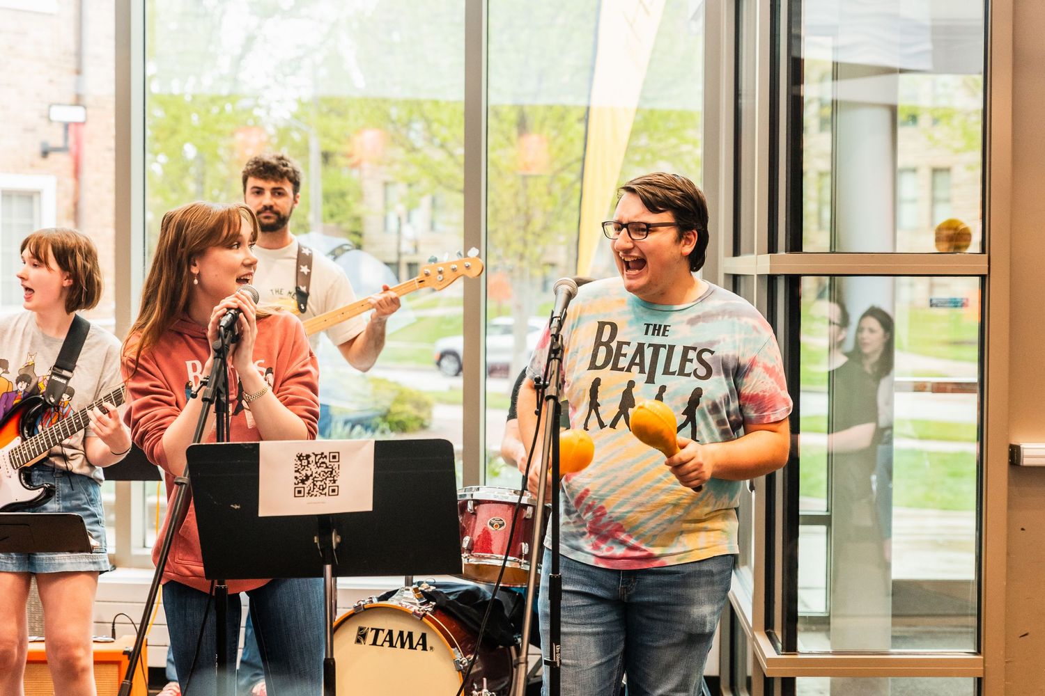 Student band performs Beatles cover songs in a casual indoor venue with guitars and microphone.