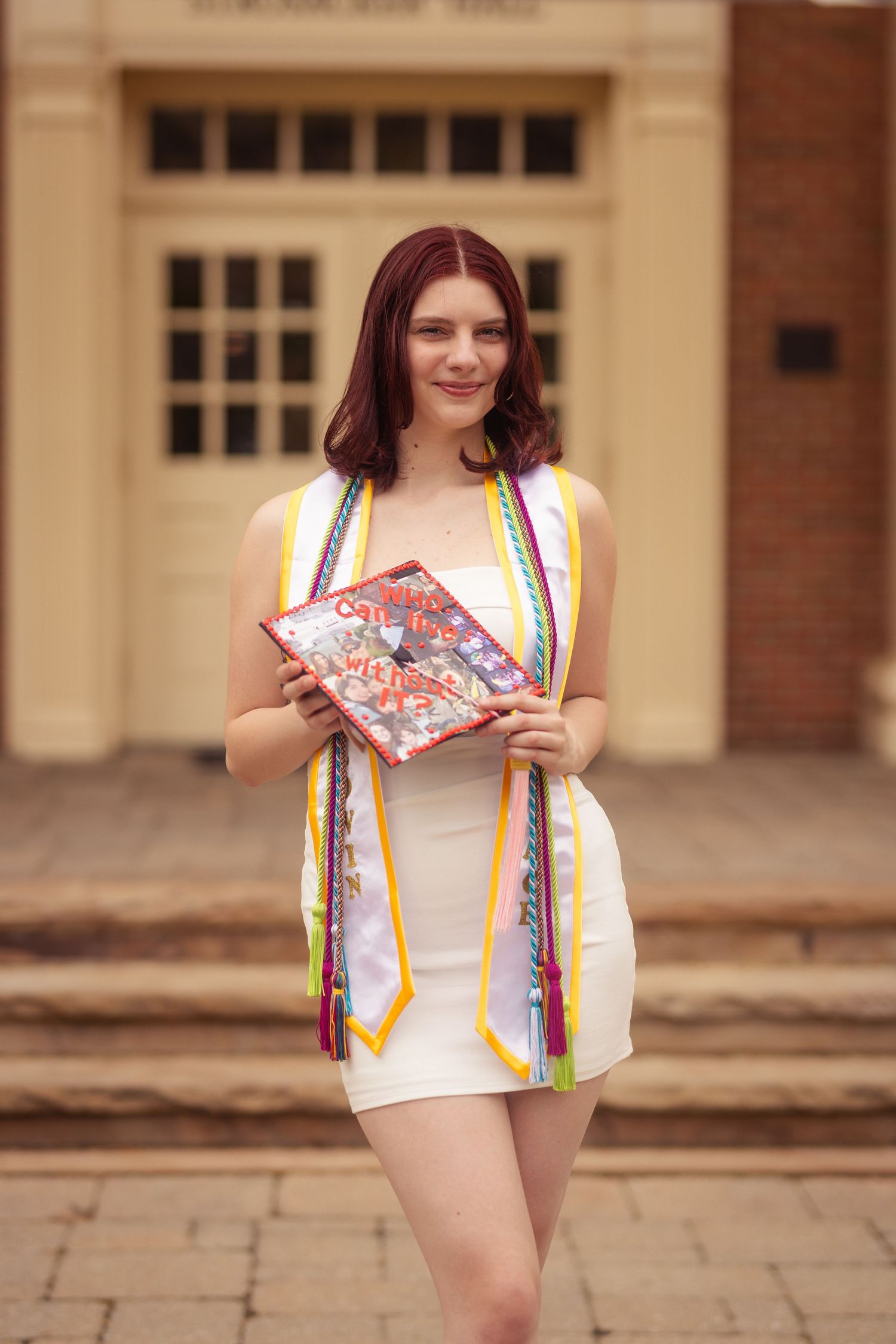 Smiling female graduate in white dress holding decorated cap with colorful honor cords.