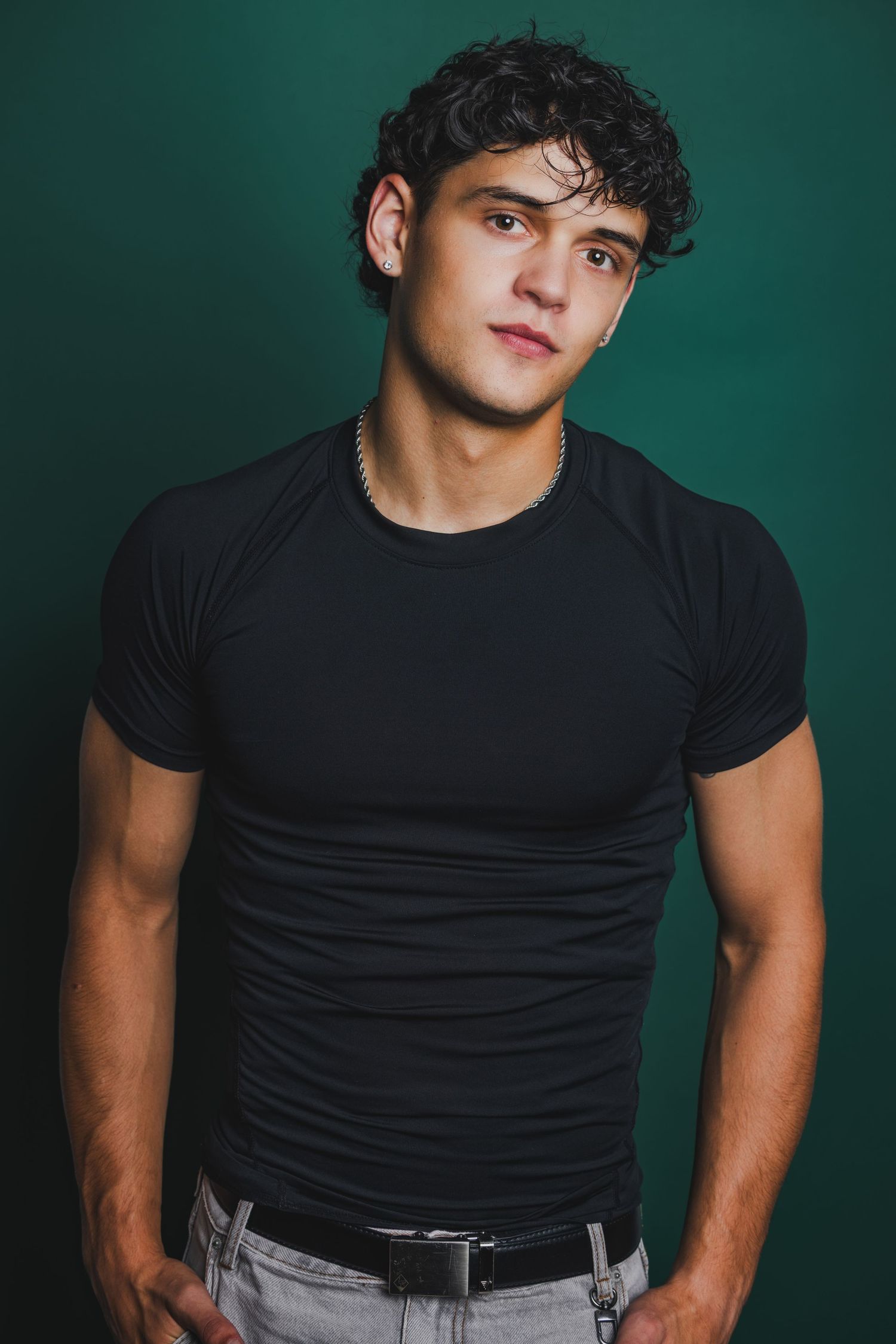 Athletic young man with curly hair in fitted black t-shirt posing against teal background.